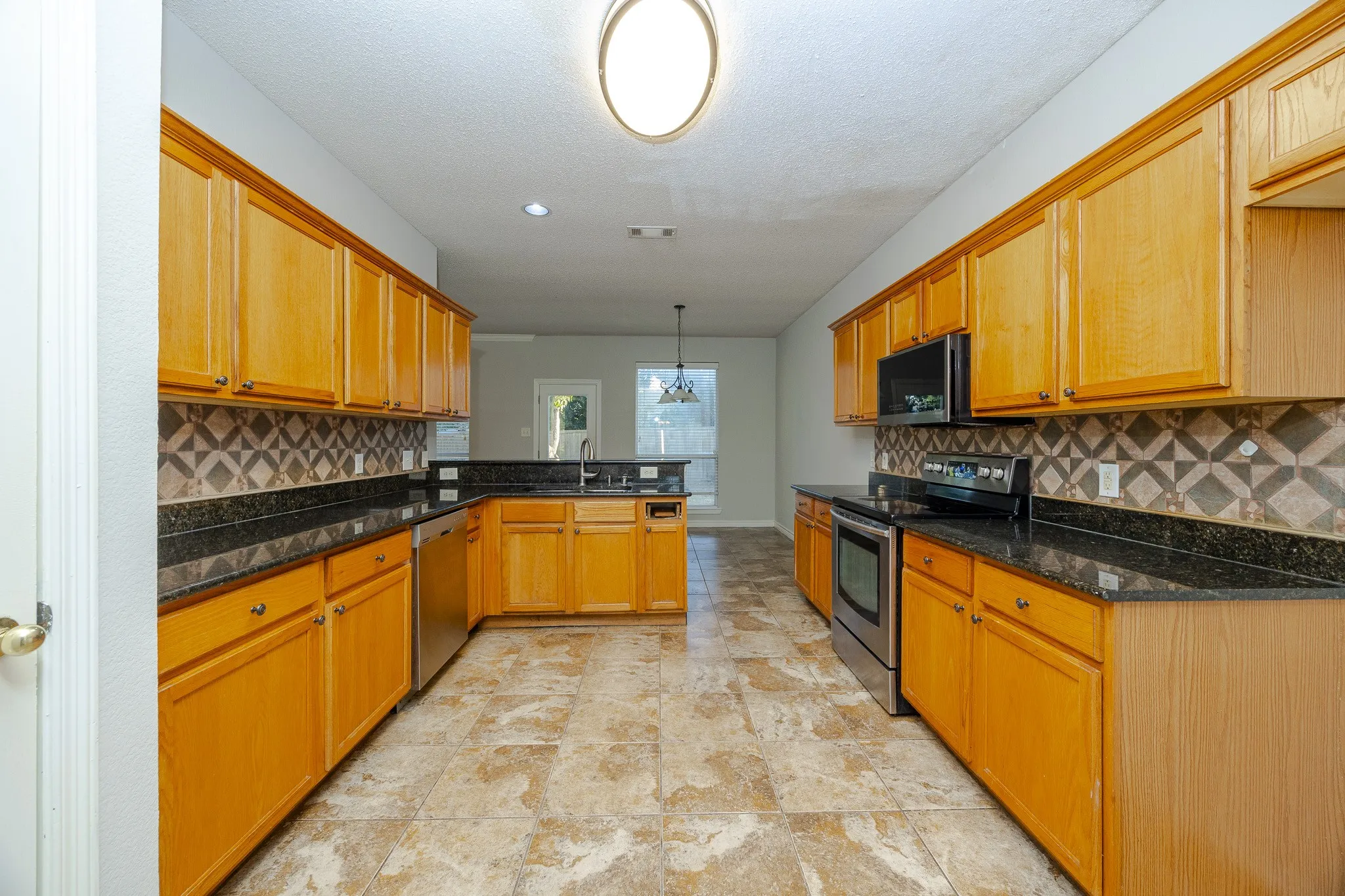 Kitchen featuring wood cabinetry, dark countertops, a patterned tile backsplash, stainless steel appliances, and large format tile flooring