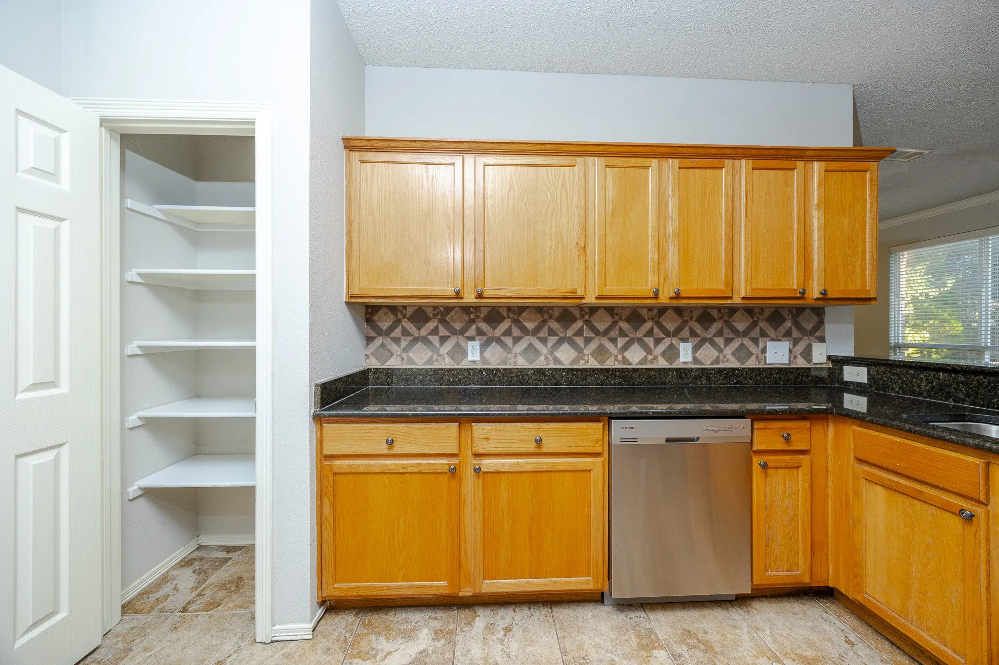 Kitchen featuring wood cabinetry, dark countertops, tiled backsplash, stainless steel dishwasher, and pantry with shelving