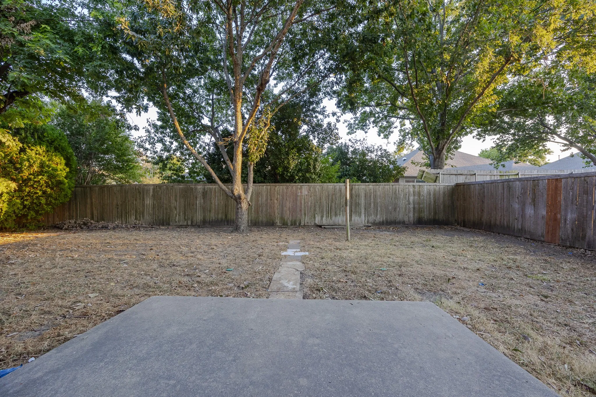 Spacious backyard featuring a concrete patio, wood fencing, mature trees, and a stone-paver walkway