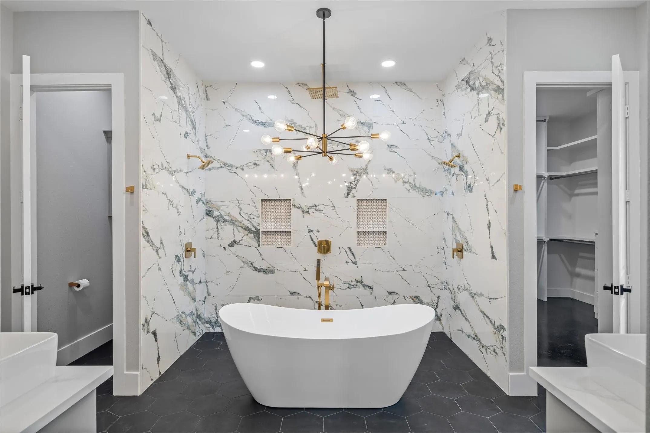 Freestanding soaking tub centered in a spacious bathroom featuring patterned wall tile, dark hexagonal floor tile, and a contemporary chandelier