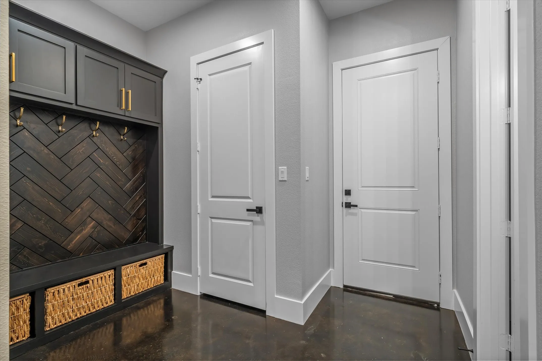 Built-in mudroom cabinetry featuring a dark wood herringbone back panel, gold-tone hardware, and integrated storage bench with woven baskets