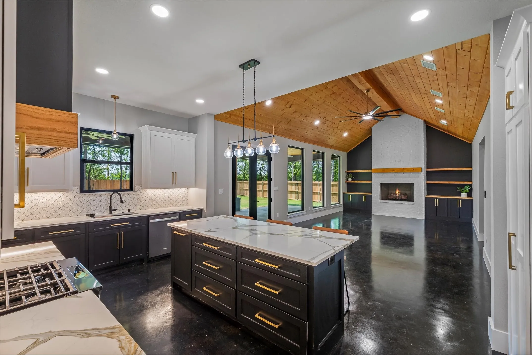 Open-concept living space featuring a vaulted wood-paneled ceiling, a central kitchen island with a quartz countertop, a gas range with a modern vent hood, a white brick fireplace with a wood mantel, and built-in shelving and cabinetry