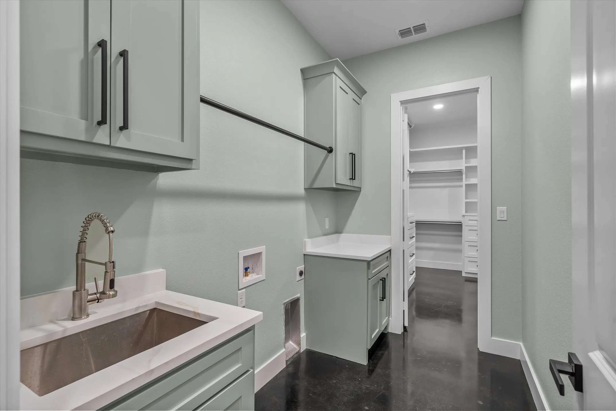 Laundry area featuring light green cabinetry with matte black hardware, a stainless steel sink with a gooseneck faucet, white solid surface countertops, and a clothes hanging rod