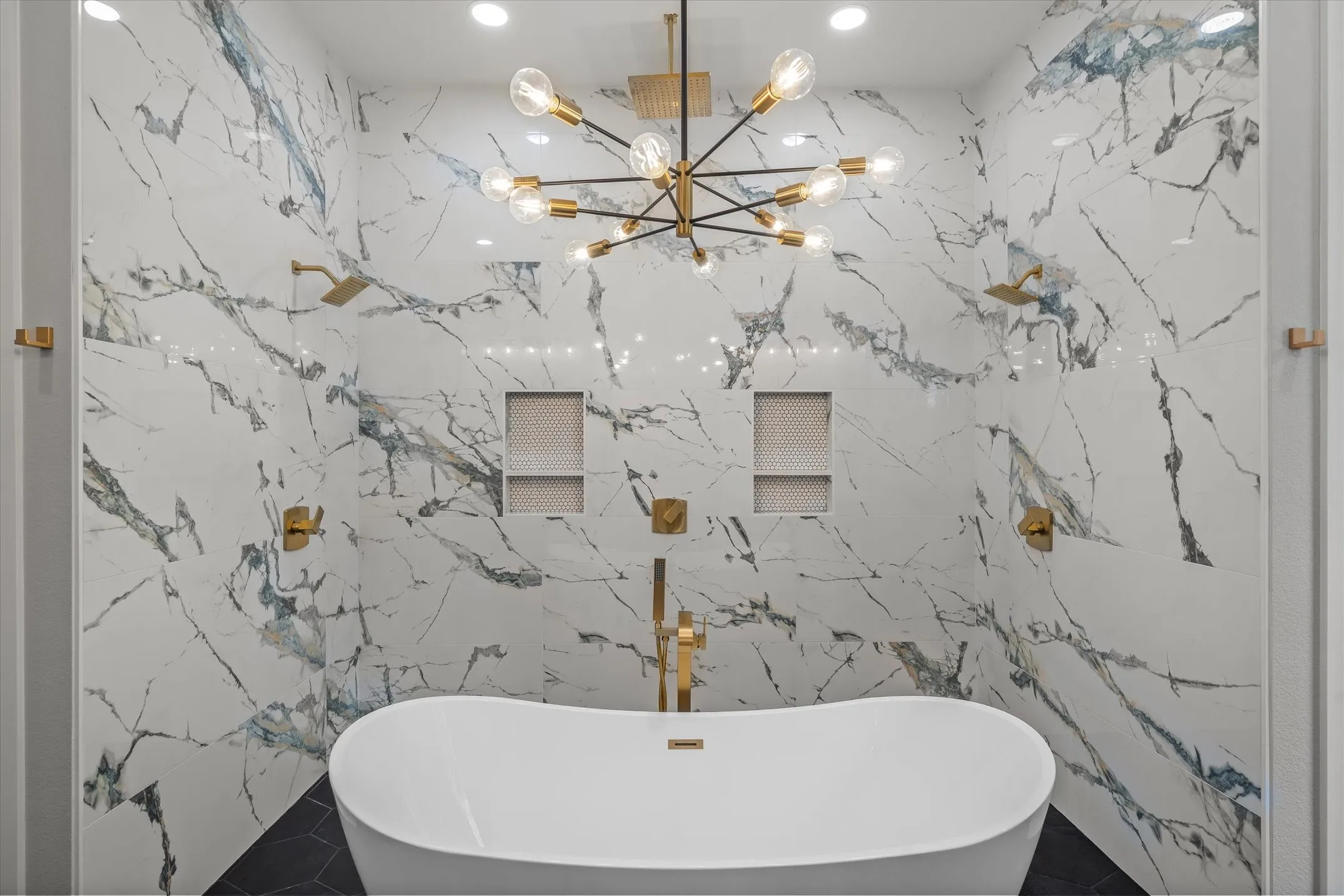 Freestanding soaking tub centered within a spacious wet room featuring large format marble-look tile walls, dual gold-finish rain shower heads, and built-in tiled shower niches