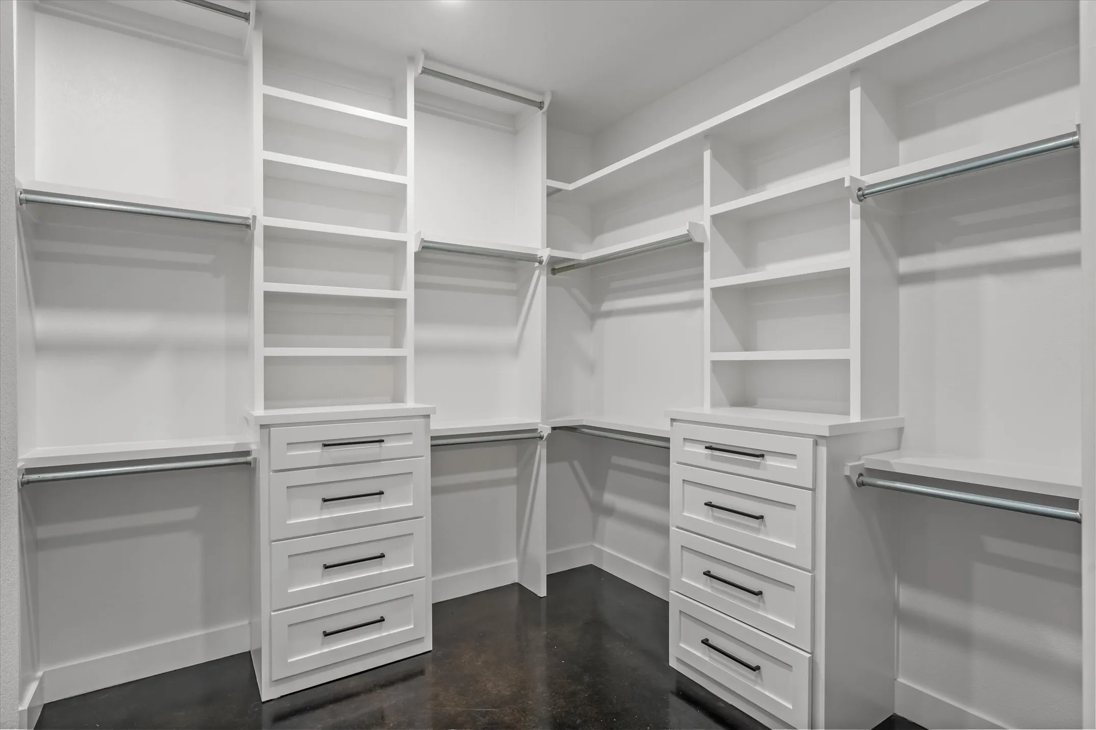 Custom closet system featuring white cabinetry and shelving, built-in drawer units with matte black hardware, multiple hanging rods, and dark flooring