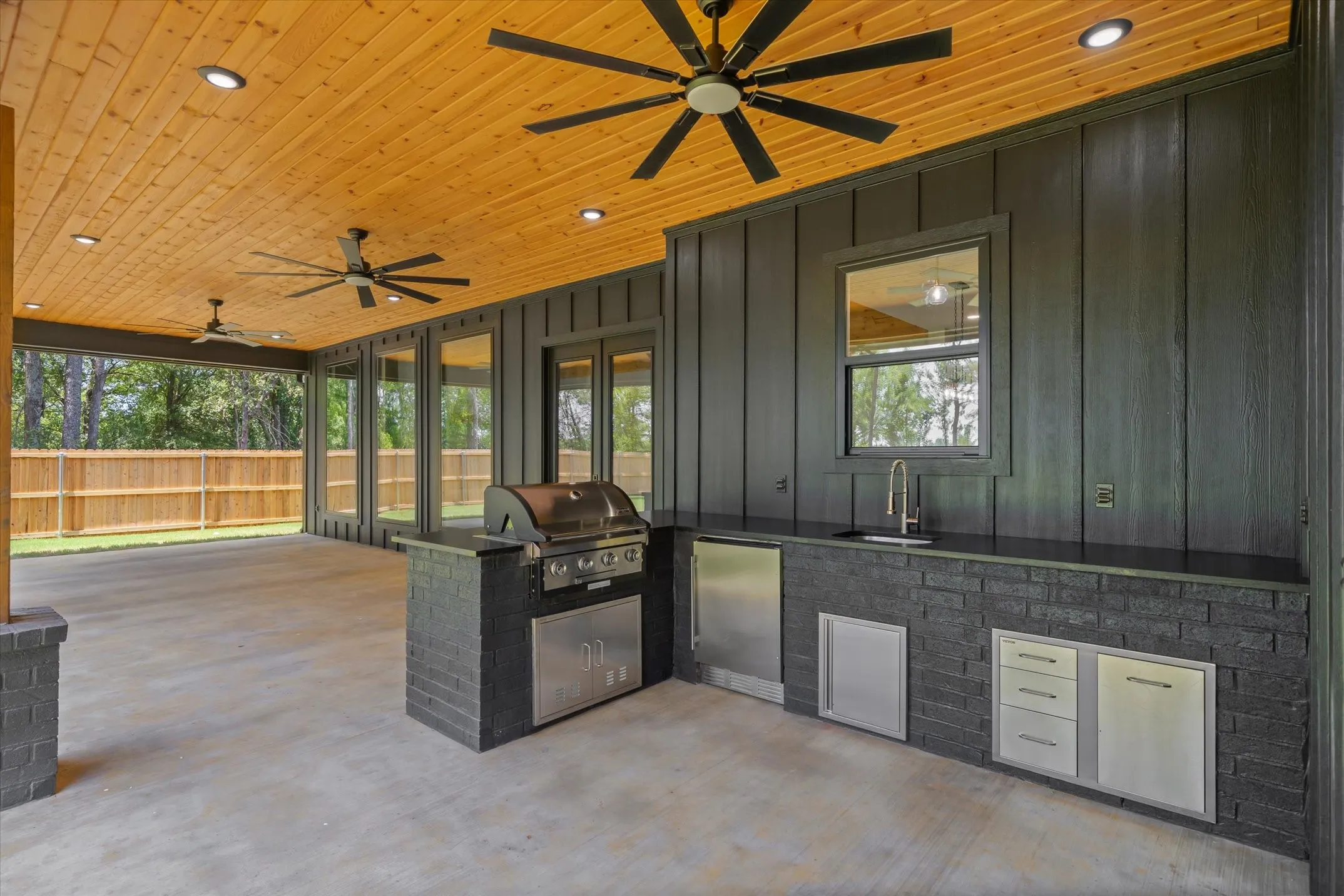 Spacious covered patio featuring a wood-panel ceiling with recessed lighting, an outdoor kitchen with stainless steel appliances, a built-in grill, and a black brick island with a sink and countertop