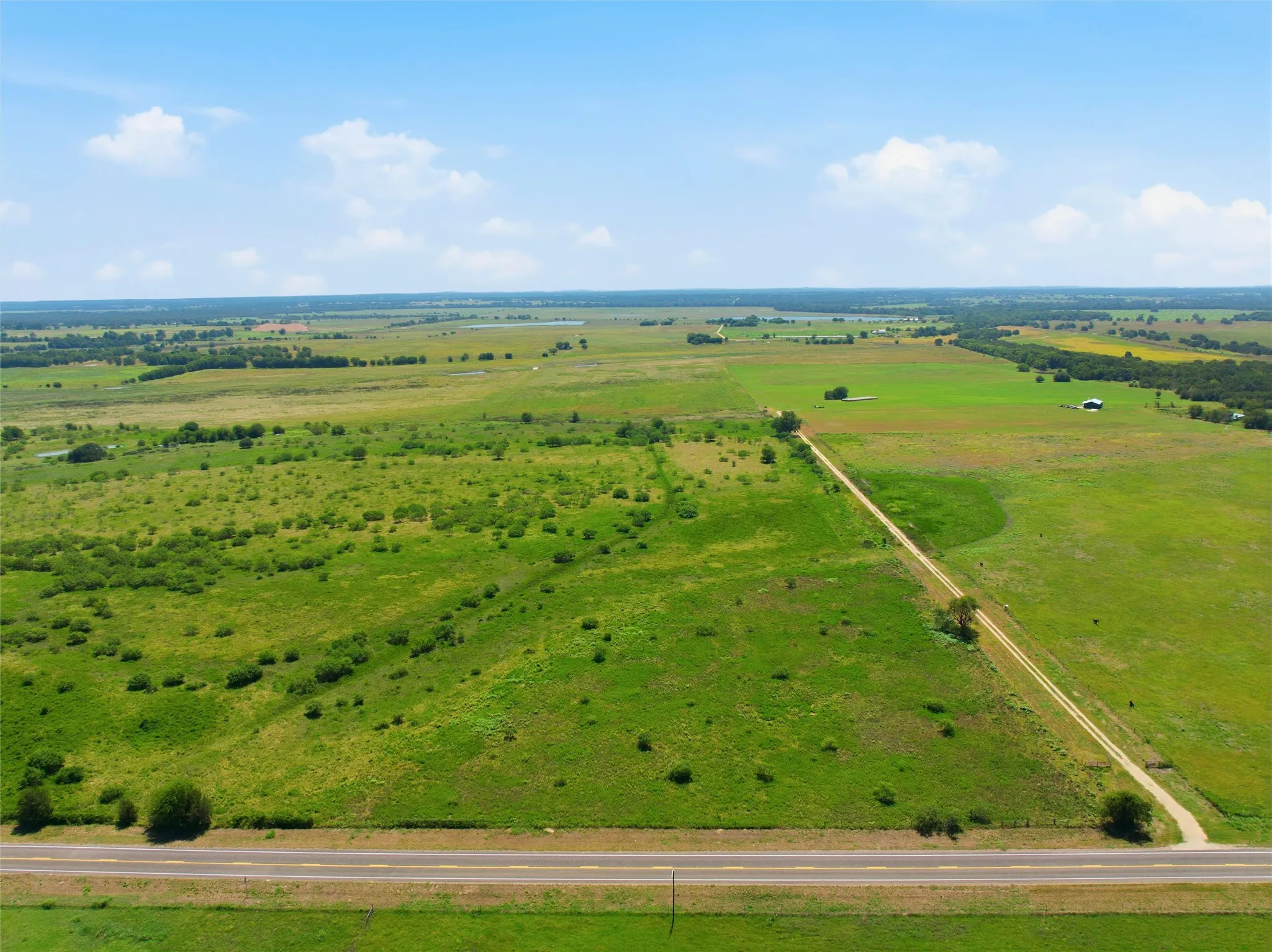 Expansive rural landscape featuring a paved road bordering open fields, a long dirt path extending into the property, and scattered natural vegetation