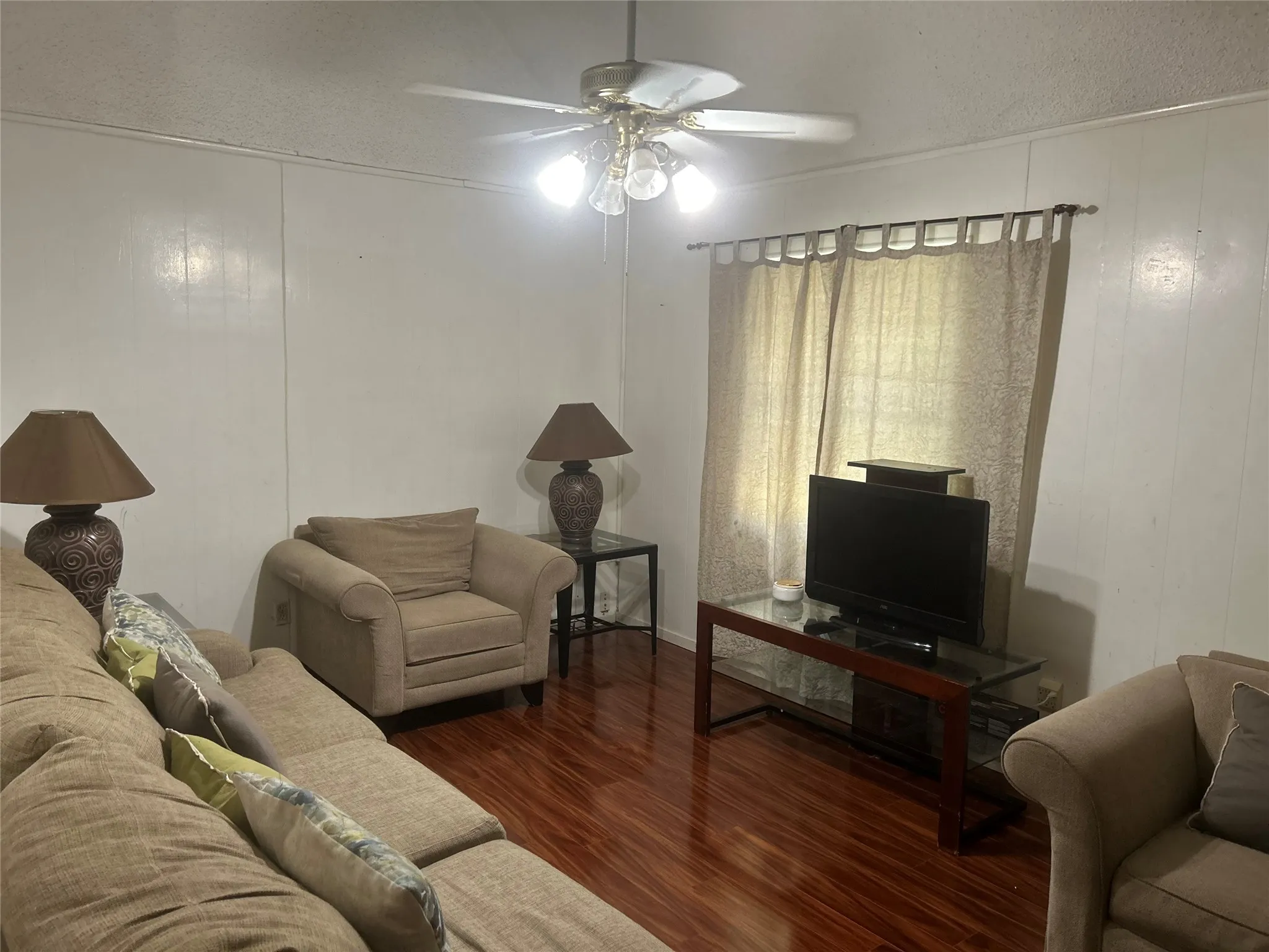 Spacious living area featuring wood-finish flooring, white panel walls, and a ceiling fan with integrated lighting