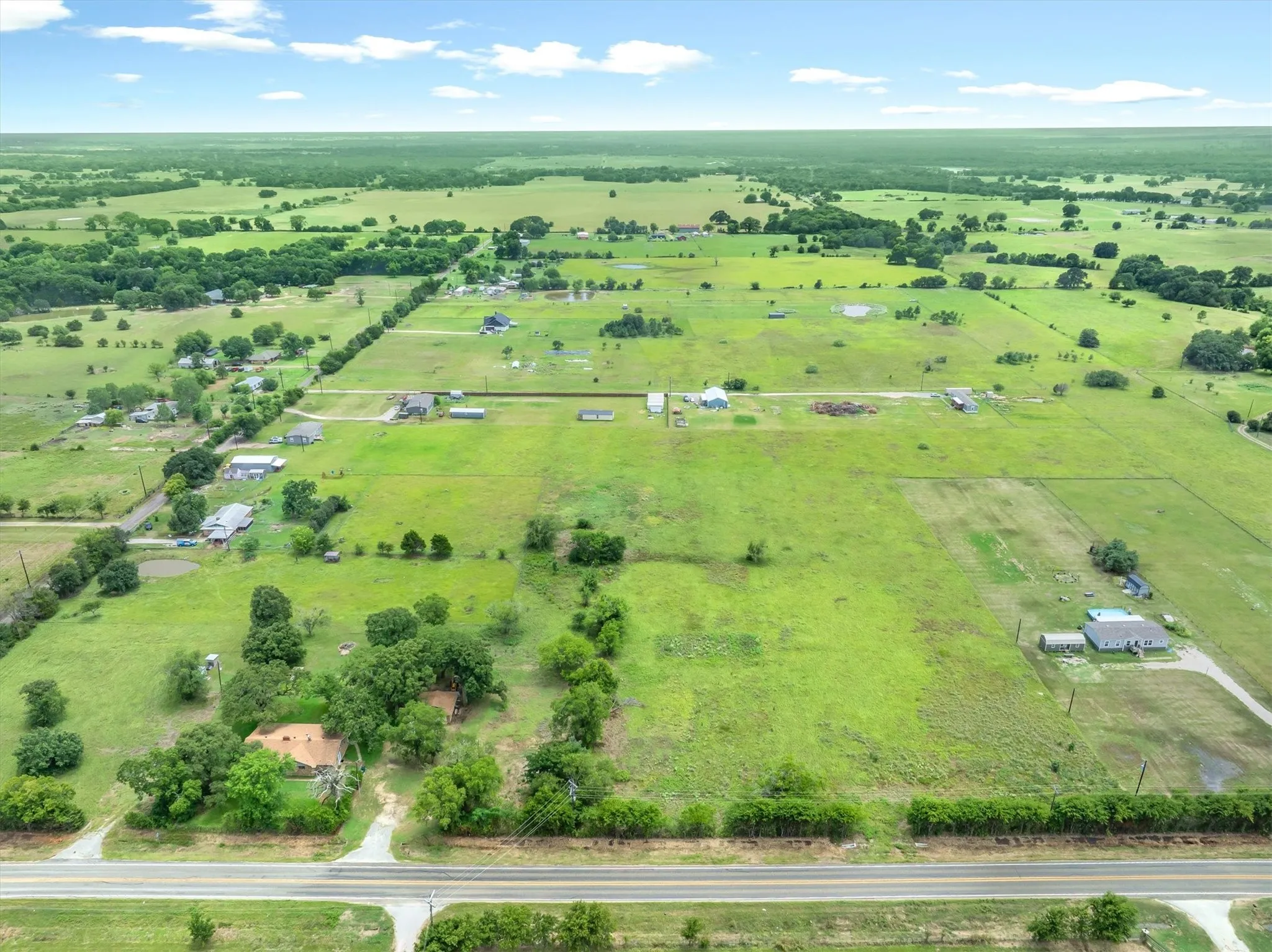 Expansive rural landscape featuring a single-story residence with a tan roof, mature trees, and a paved road frontage
