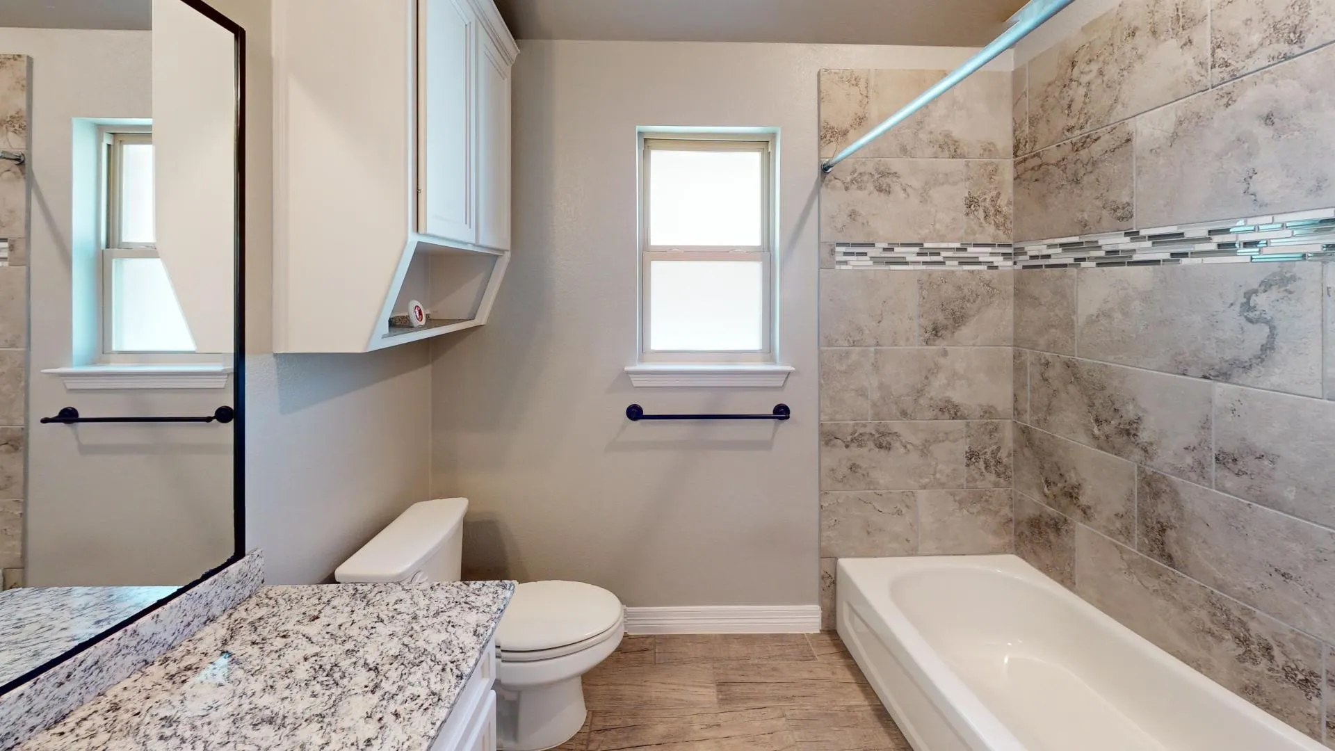 Full bathroom featuring a single window, white bathtub with a tiled surround, decorative glass tile accent band, wood-finish flooring, and a vanity with a speckled countertop