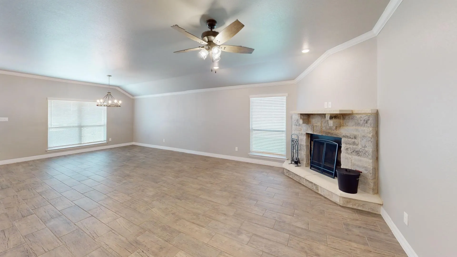 Spacious living area featuring wood-finish tile flooring, a stone-surround fireplace, vaulted ceilings, a ceiling fan, and multiple windows with blinds