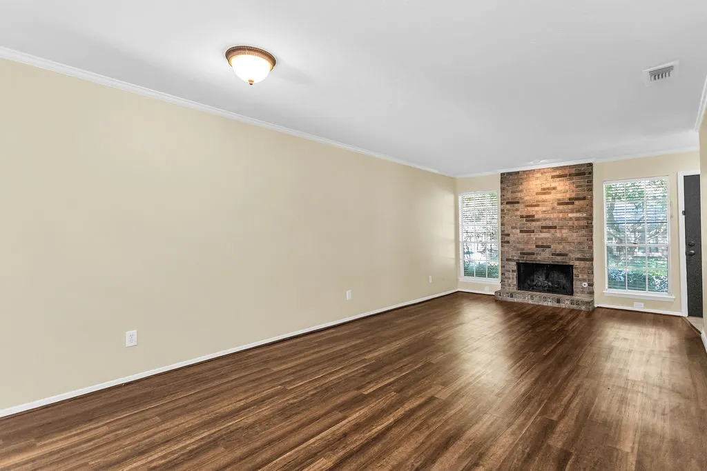 Spacious living area featuring wood-finish flooring, a brick fireplace, two large windows with blinds, crown molding, and a flush mount light fixture