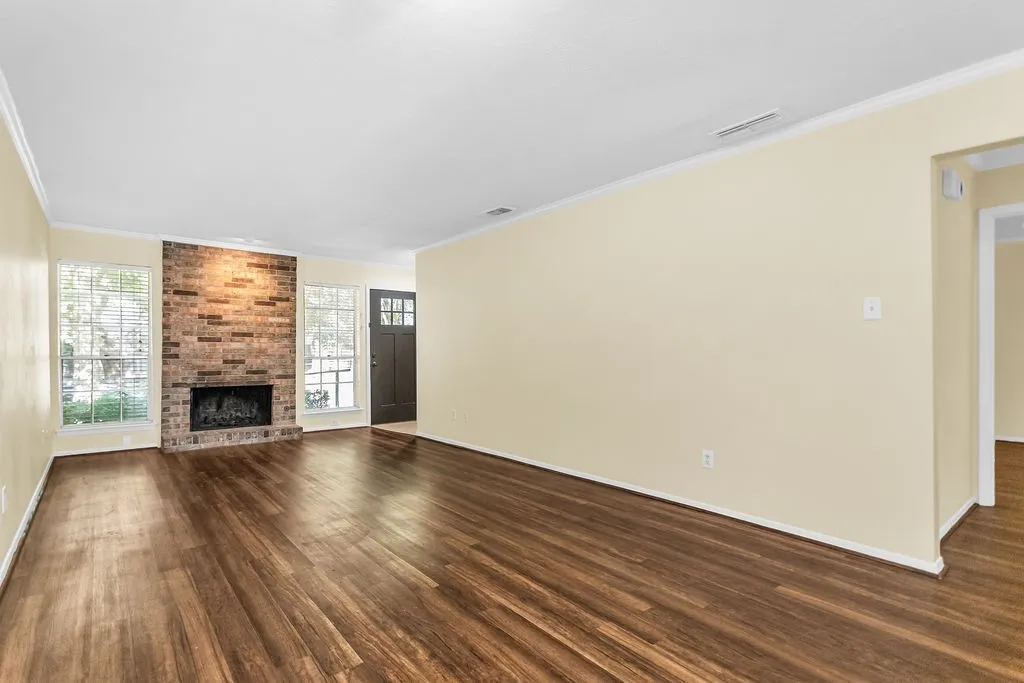 Expansive living area featuring wood-finish flooring, a brick-surround fireplace, multiple large windows, and a paneled front door with decorative glass inserts