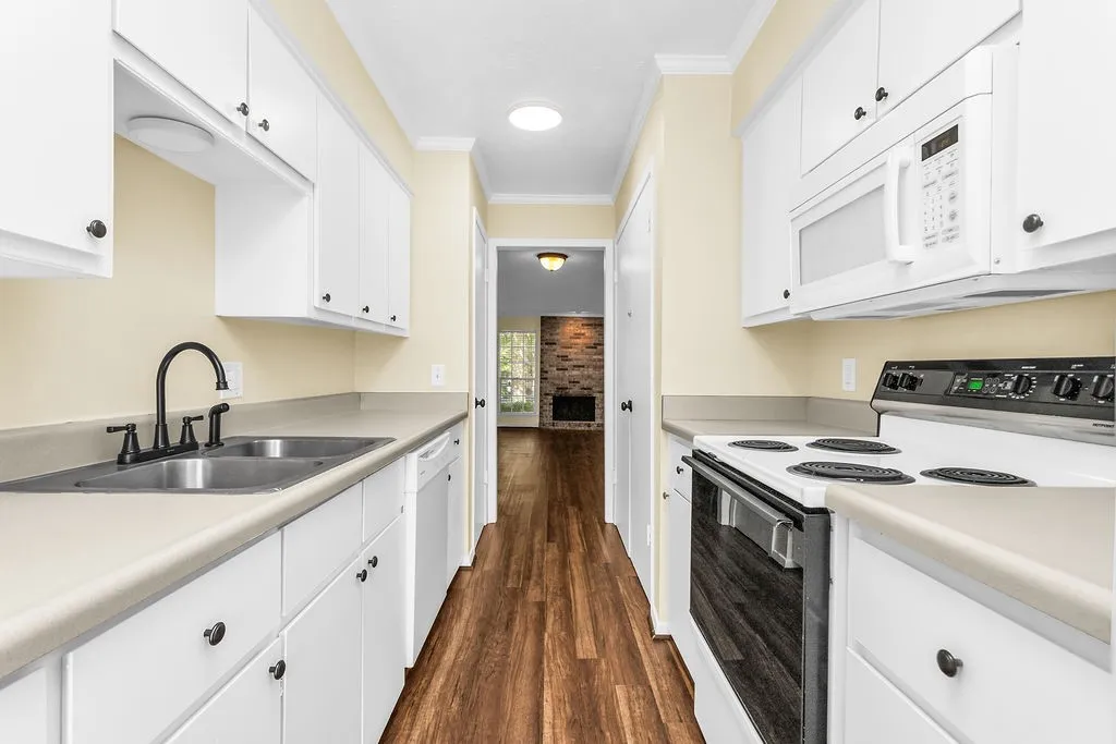 Galley kitchen featuring white cabinetry, light-toned countertops, a stainless steel double sink with a matte black faucet, and wood-finish flooring