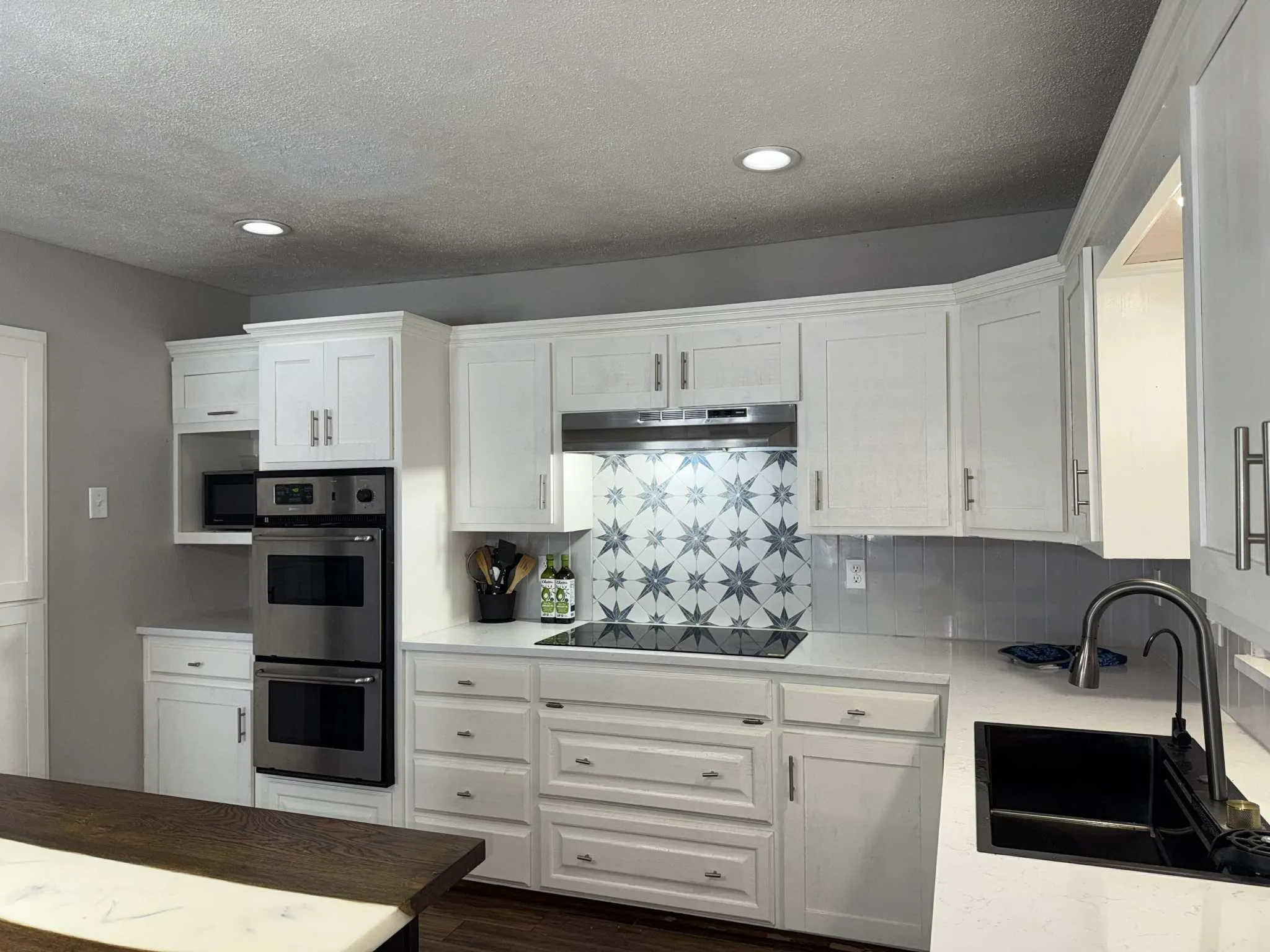 Kitchen featuring white cabinetry, light-toned countertops, a black composite sink, and a star-patterned backsplash above a built-in cooktop