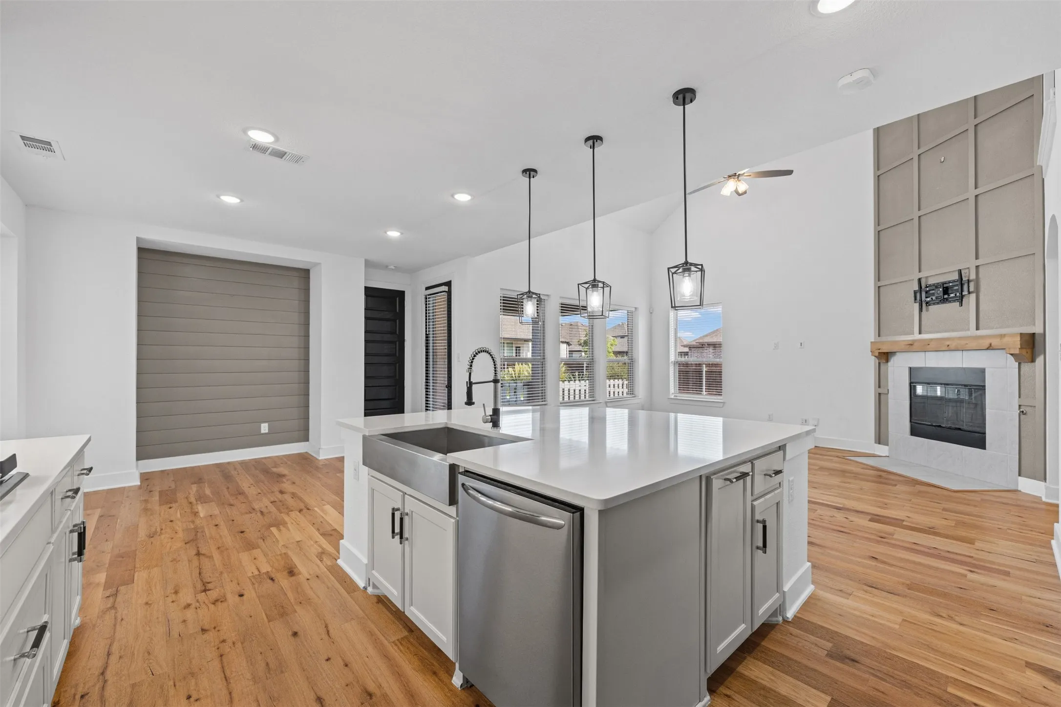 Spacious open-concept interior featuring light wood-finish flooring, white cabinetry, a large kitchen island with a farmhouse sink, and a fireplace with a wood mantle and tile surround