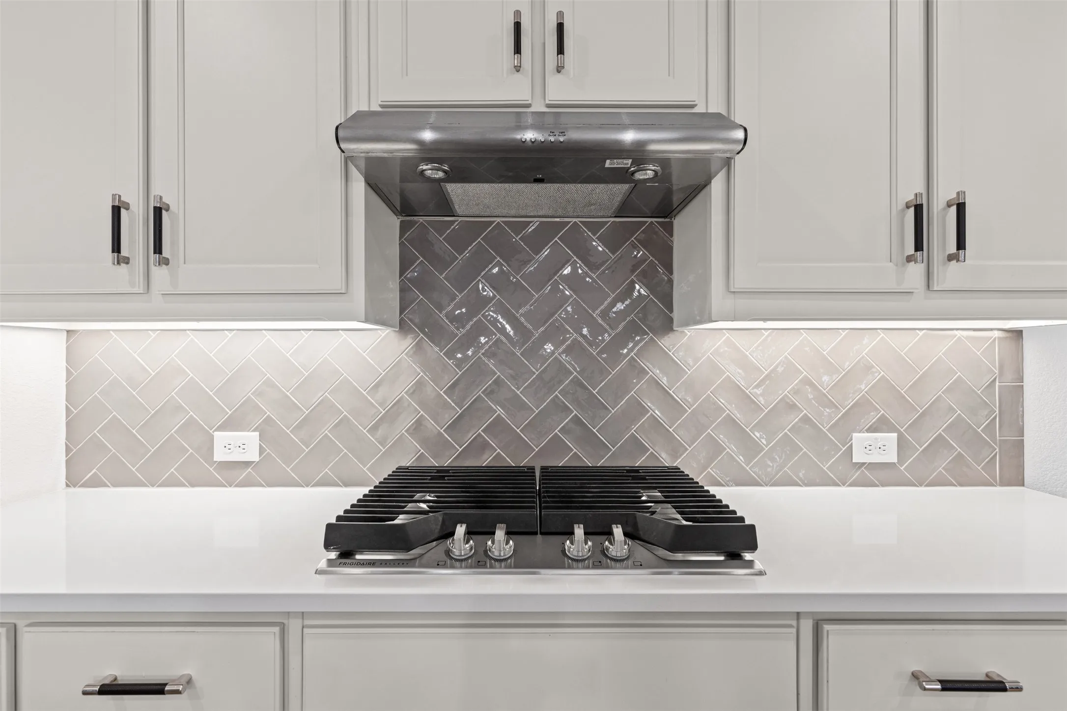 White countertops, stainless steel gas cooktop, stainless steel range hood, light gray shaker cabinets, and herringbone tile backsplash