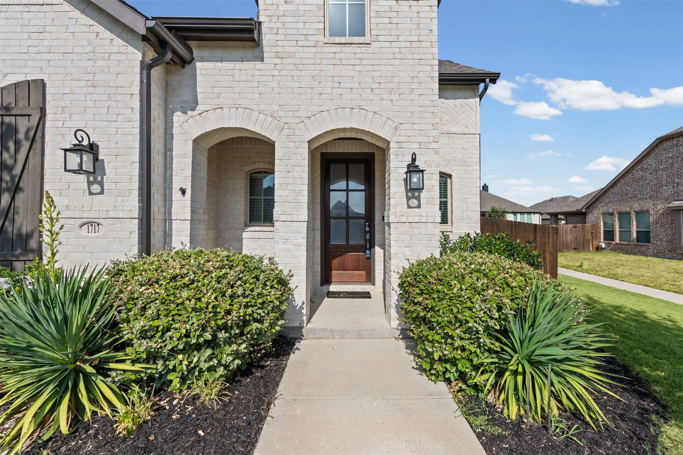 Light brick exterior with dark wood shutters, arched entryway, dark wood entry door with glass panels, and built-in exterior lighting