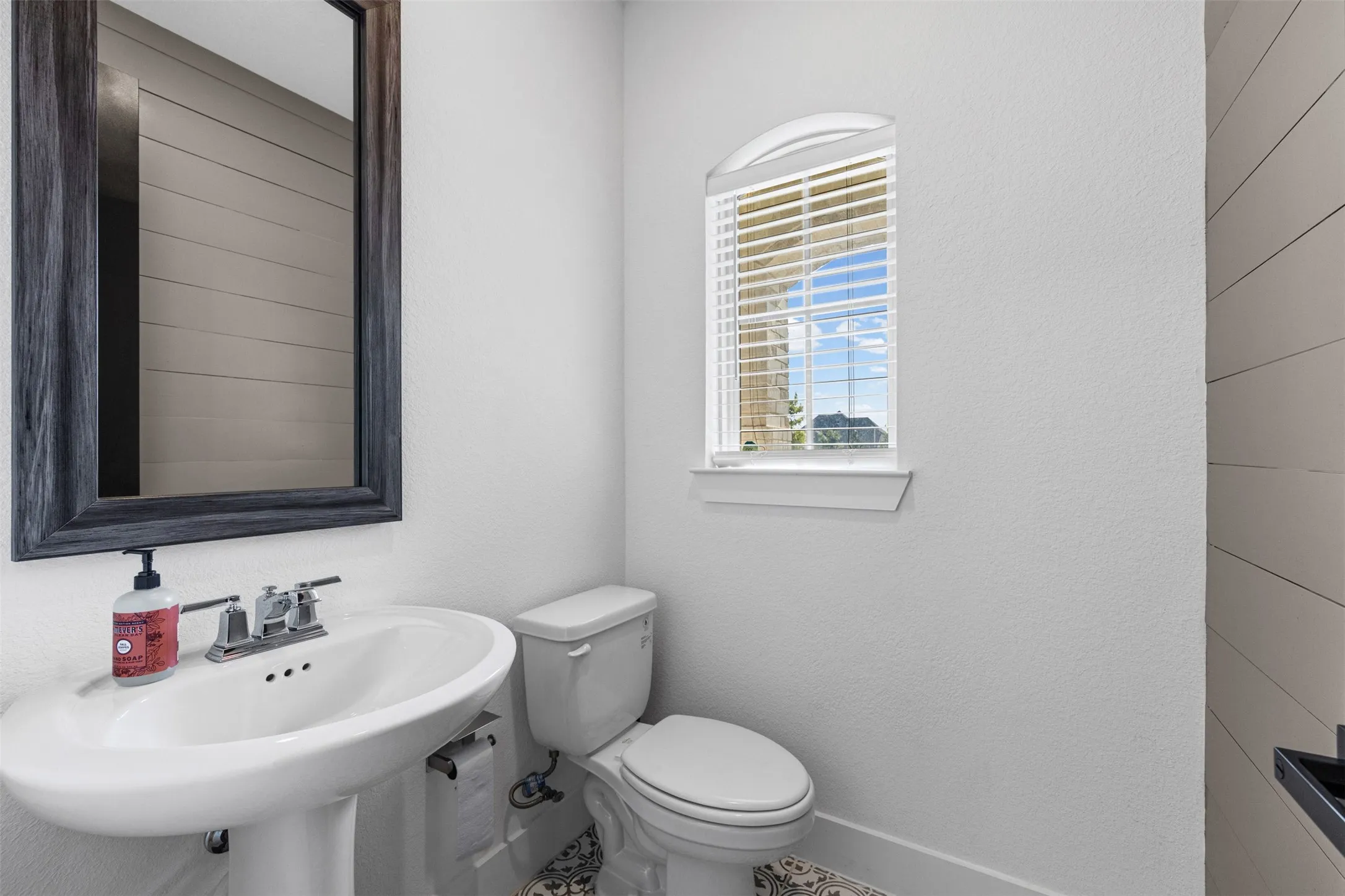 Pedestal sink with chrome faucet, arched window, decorative floor tiling, and shiplap wall accent