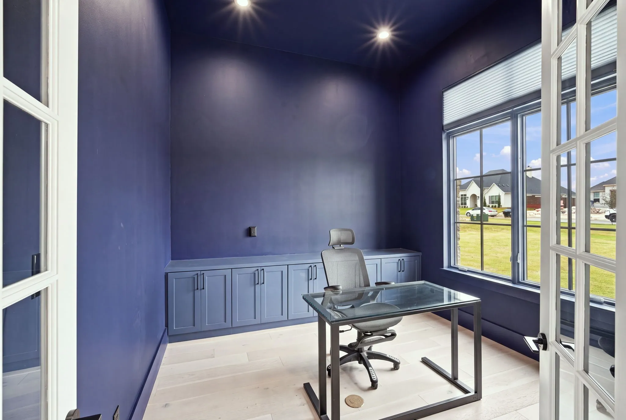 Modern home office featuring deep blue accent walls, recessed ceiling lighting, light wood-finish flooring, built-in cabinetry with multiple storage compartments, and expansive windows with natural light and outdoor landscape visibility