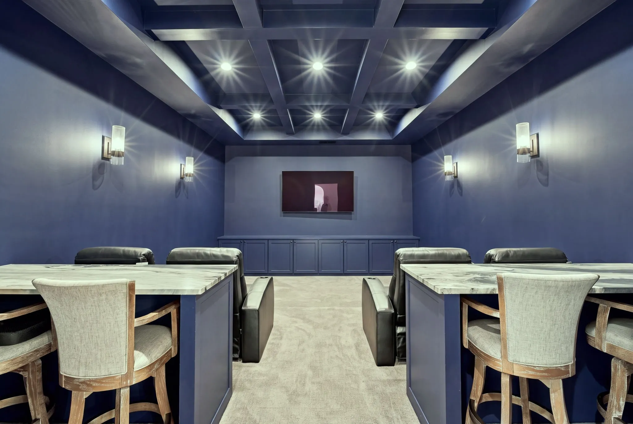 Dedicated home theater featuring tiered seating with upholstered benches, recessed ceiling with integrated lighting design, blue-painted walls, wall-mounted sconce lighting, and a projection screen