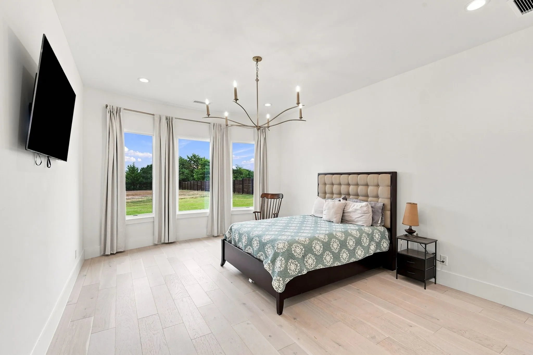 Bright primary bedroom featuring triple glass doors opening to manicured grounds, light wood-finish flooring, a modern sputnik-style chandelier, and a decorative upholstered headboard with coordinating bedding