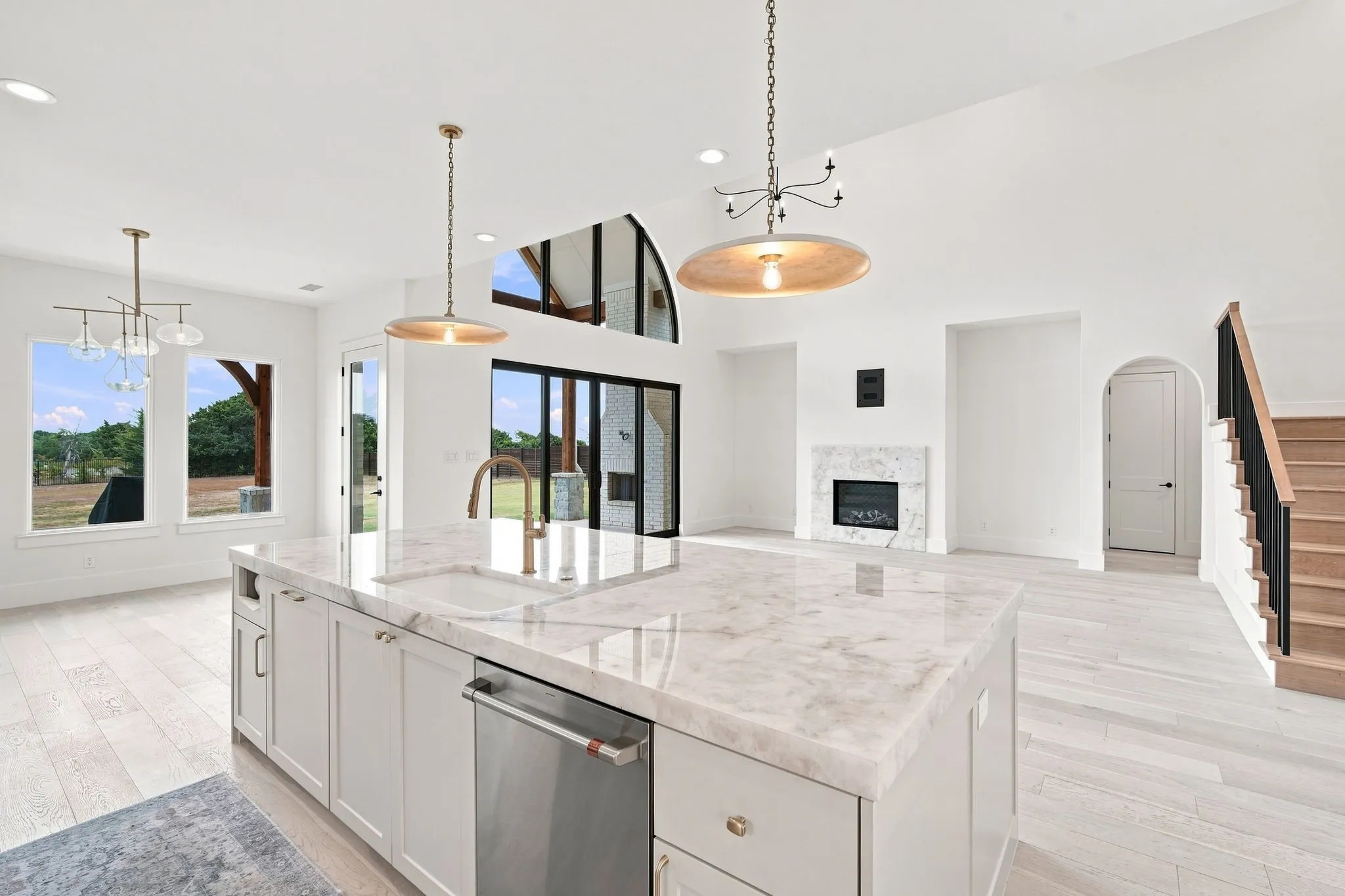 Open-concept kitchen and great room featuring a large marble-top island with waterfall edge, stainless steel appliances, white cabinetry, light wood-finish flooring, and pendant lighting