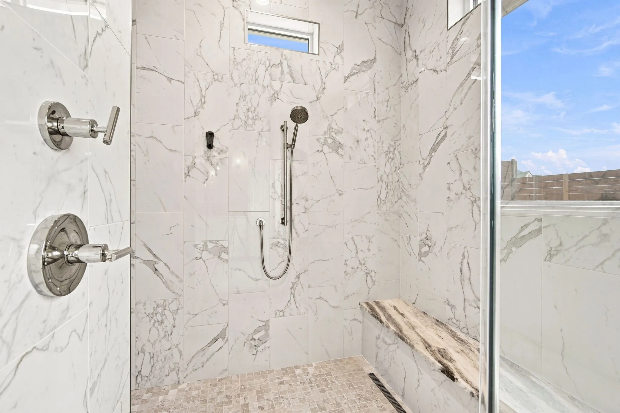 Spa-inspired shower enclosure featuring marble-look porcelain tile walls, a pebble stone floor, dual showerheads with handheld wand, and chrome fixtures throughout