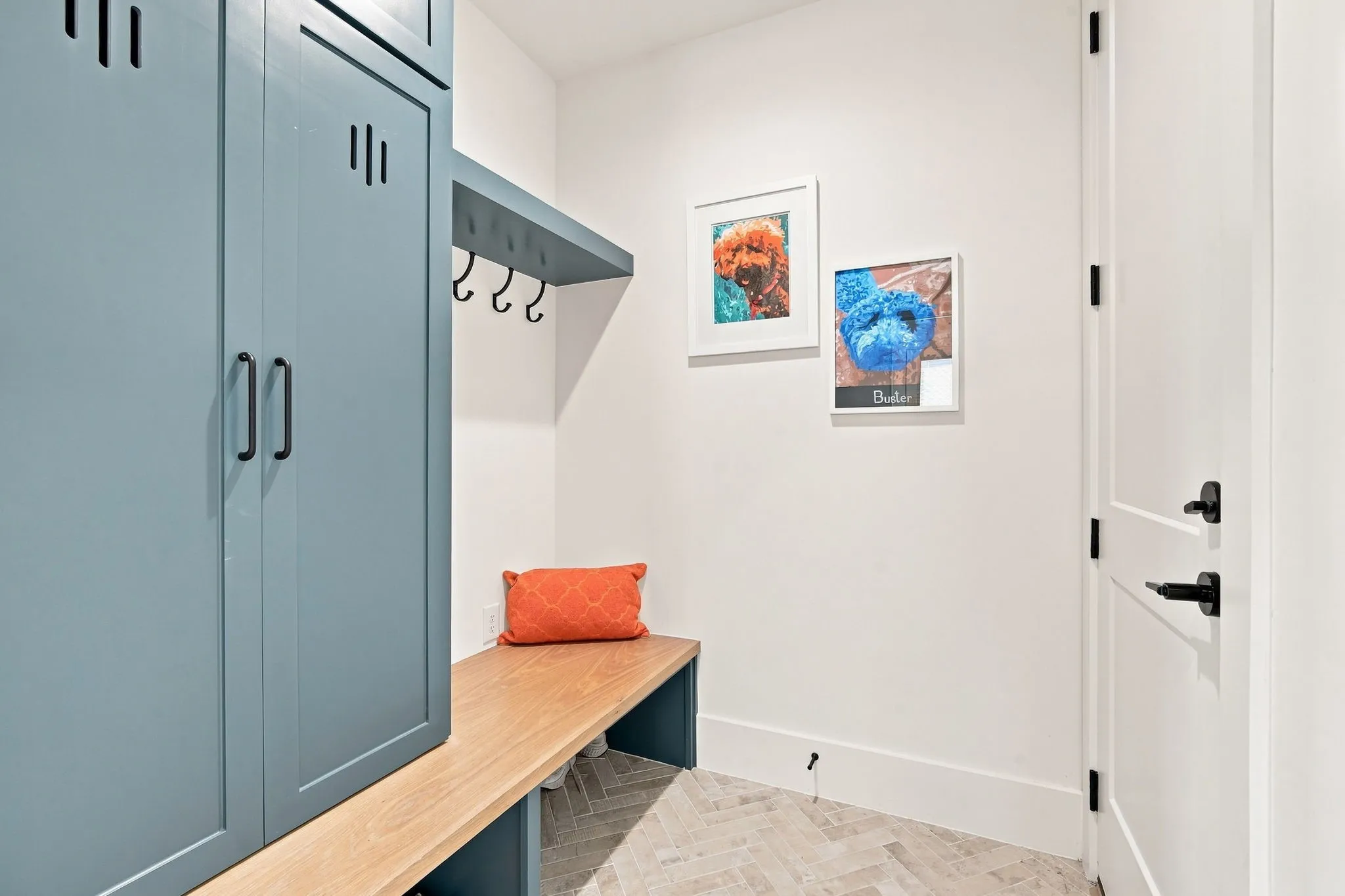 Mudroom featuring custom blue cabinetry with integrated hooks, a wood-finish built-in bench with storage, neutral tile flooring, and white painted walls accented with framed artwork