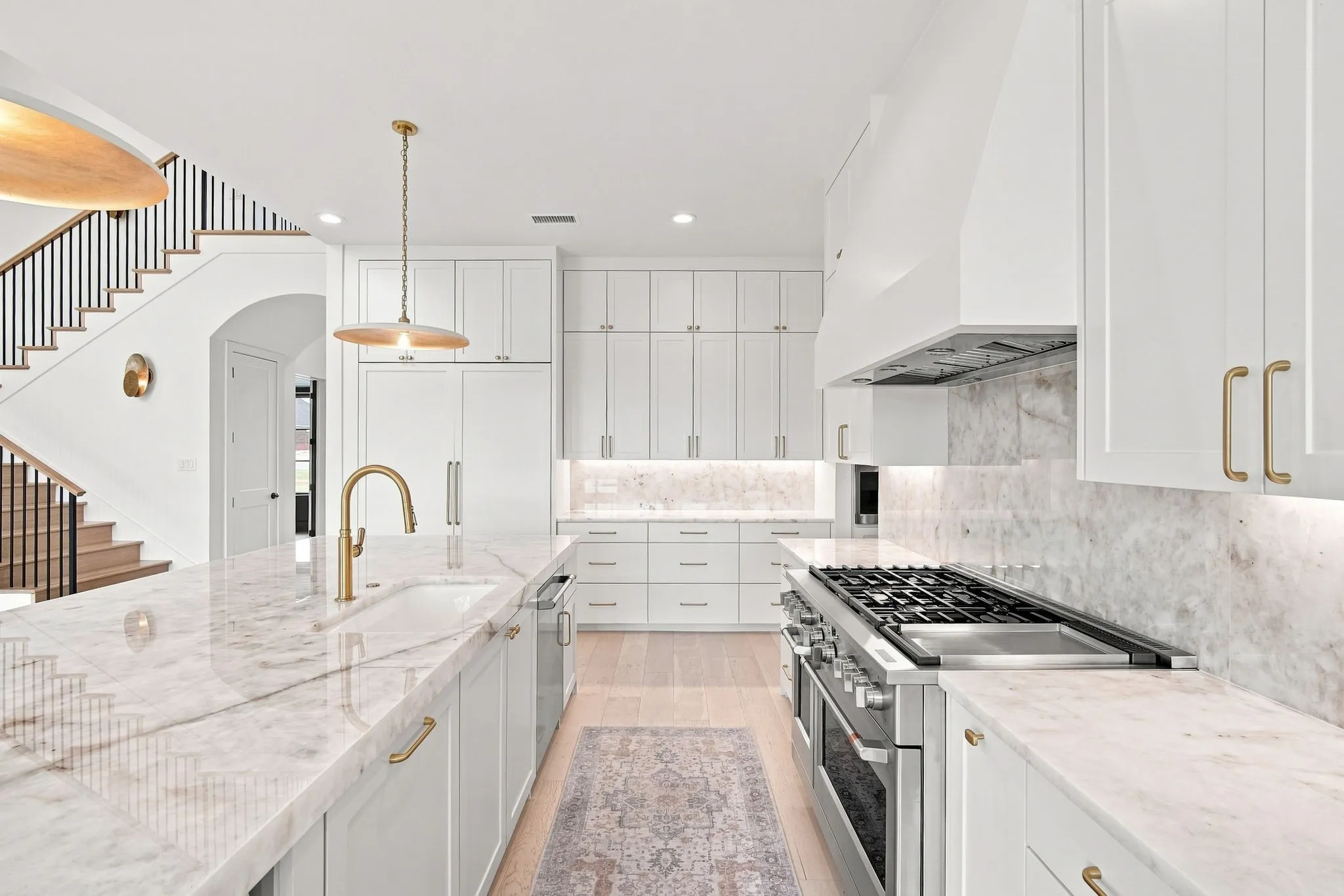 Gourmet kitchen featuring white cabinetry with brass hardware, marble countertops and backsplash, a professional-grade gas range with stainless steel hood, and a matching marble-topped island with integrated sink and brass faucet