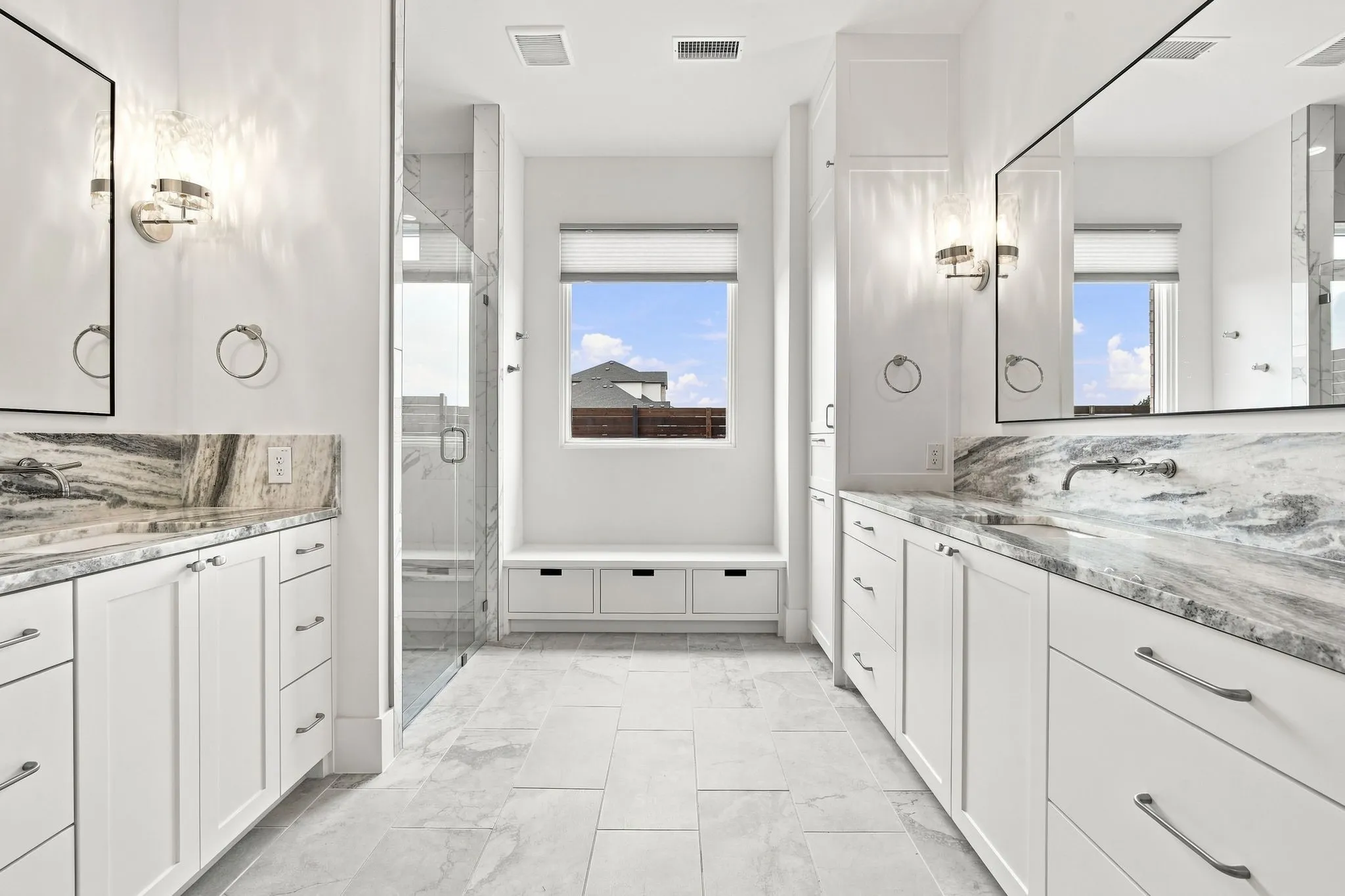 Luxurious primary bathroom suite featuring dual vanities with polished stone countertops, white cabinetry with chrome hardware, frameless glass-enclosed shower enclosures with chrome fixtures, sconce lighting, and polished stone flooring throughout