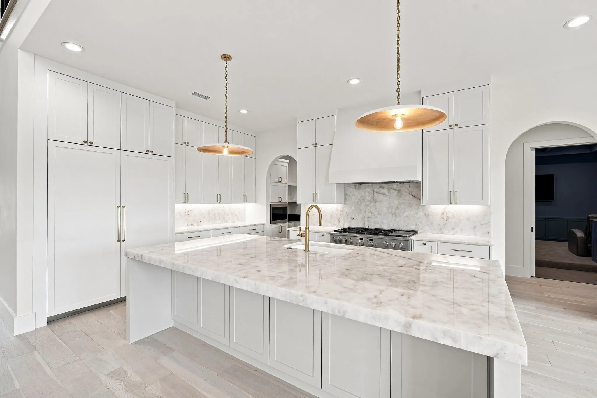 Gourmet kitchen featuring white cabinetry with brushed nickel hardware, a sprawling light-stone countertop island with waterfall edge, stainless steel appliances including a professional-range cooktop, and pendant lighting with warm brass accents