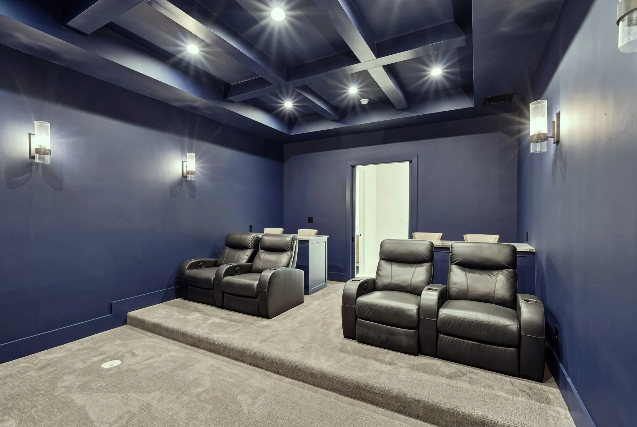 Dedicated home theater featuring tiered seating with individual recliners, deep charcoal upholstered seating, coffered ceiling with integrated recessed lighting, blue-gray wall paneling, and polished concrete flooring