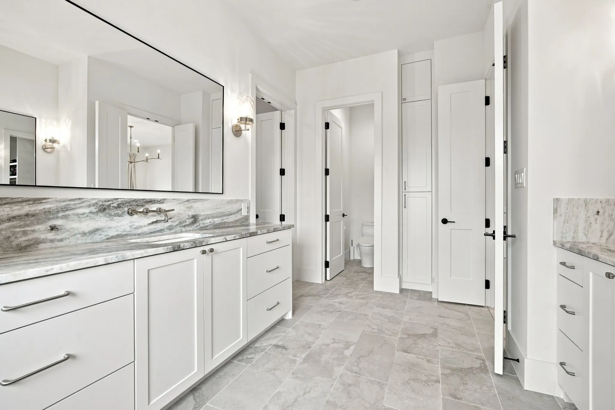 Primary Bathroom featuring dual vanities with white cabinetry, marble-finish countertops with coordinating backsplash, large framed mirror with integrated lighting, and stone-finish flooring throughout