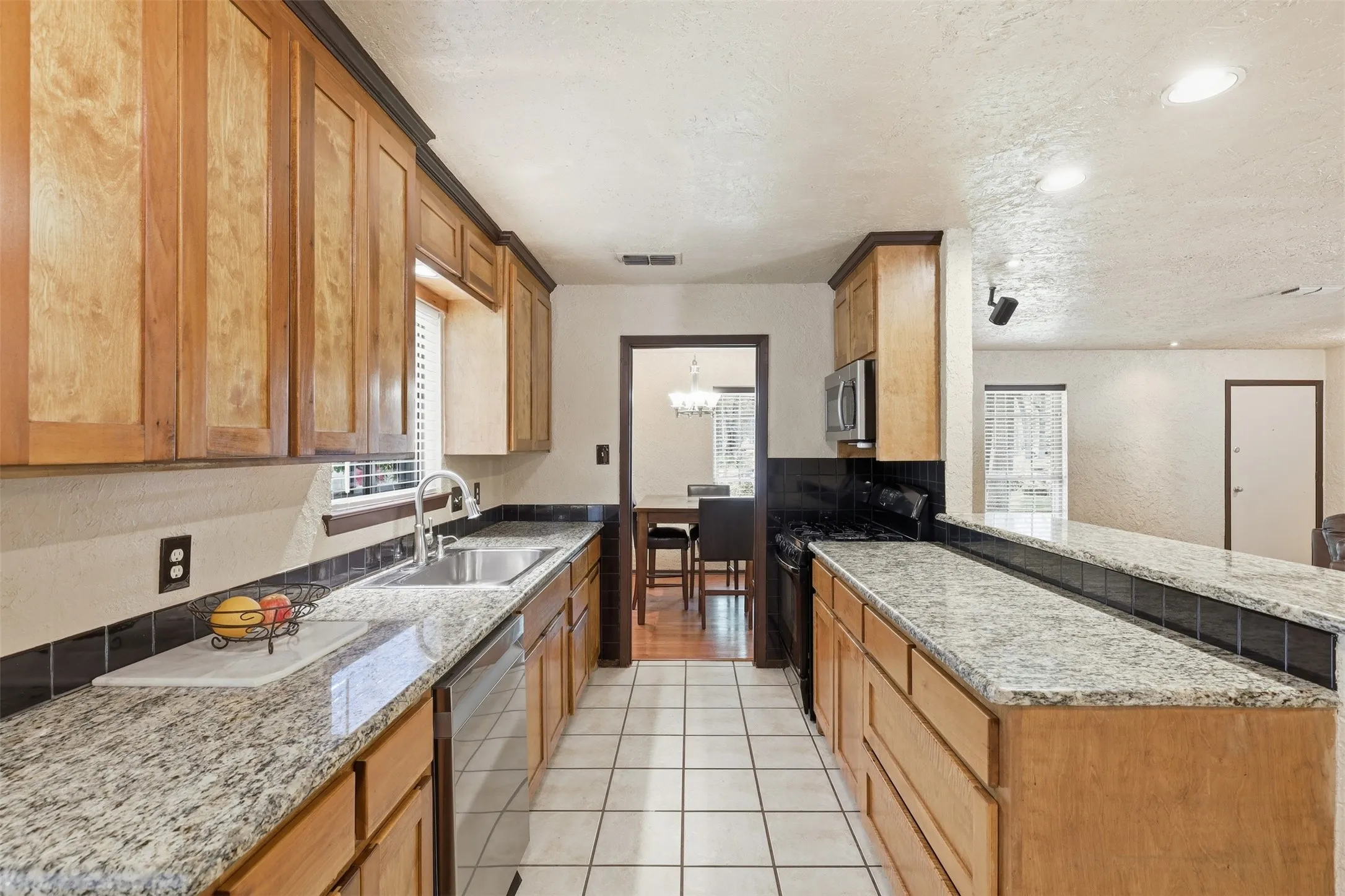Galley-style kitchen with granite countertops, wood cabinetry, double sink, tile flooring, and multiple windows.