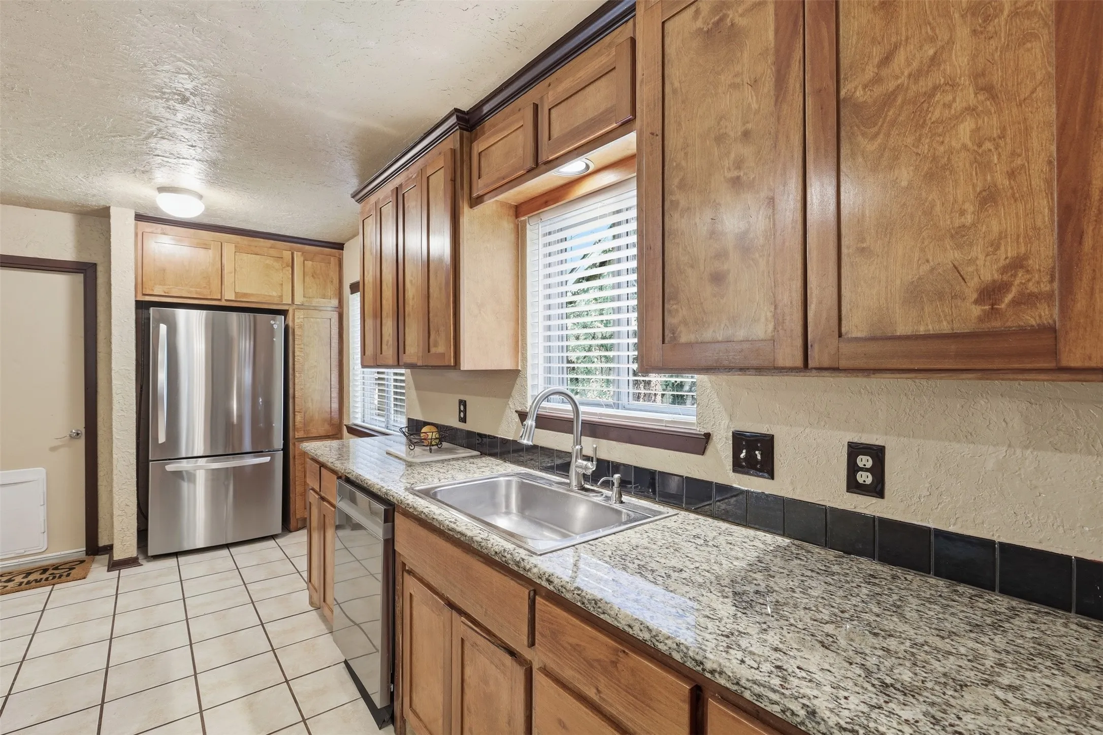 Galley-style kitchen with granite countertops, wood cabinetry, double sink, tile flooring, and multiple windows.