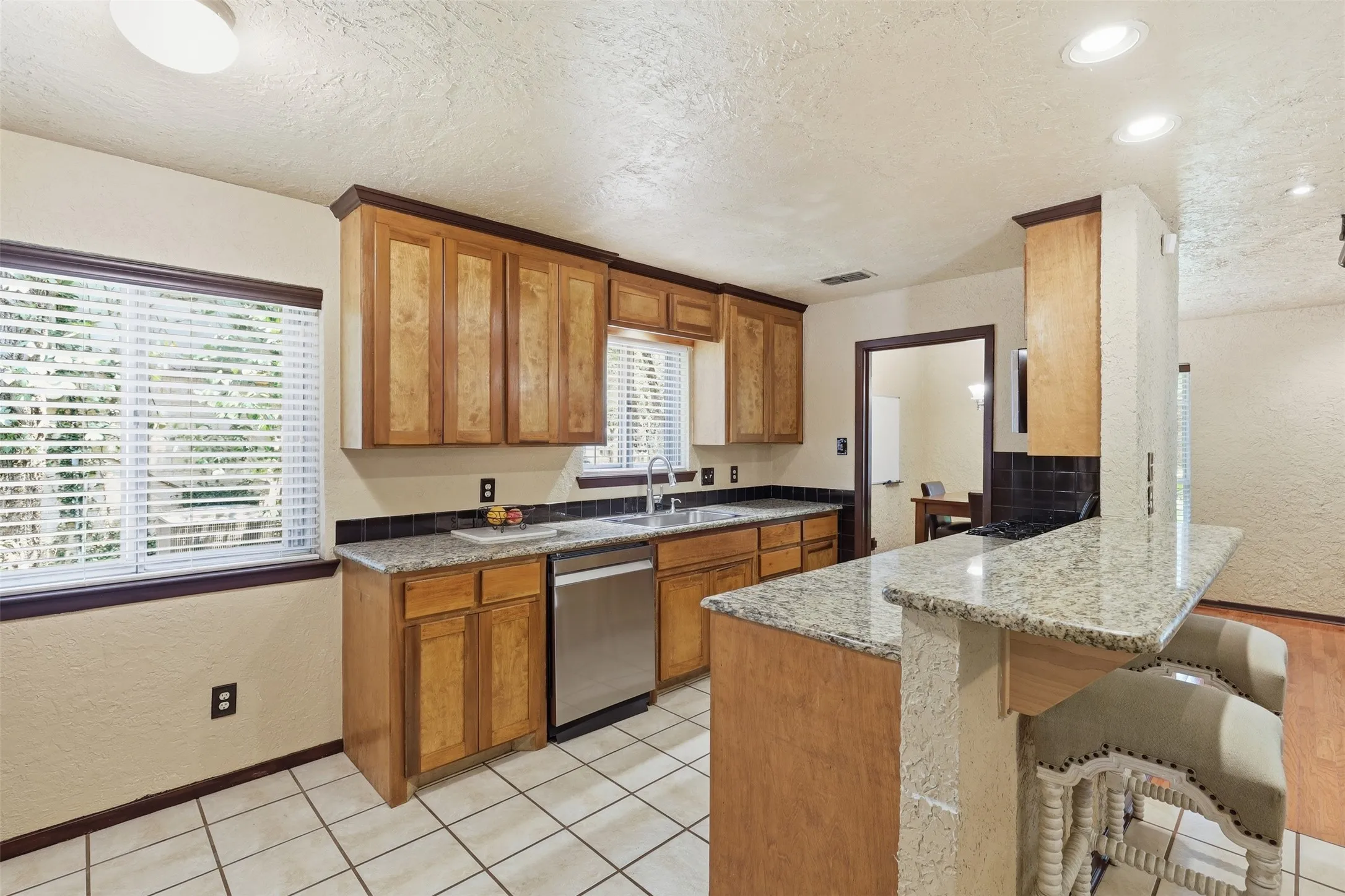 Galley-style kitchen with granite countertops, wood cabinetry, double sink, tile flooring, and multiple windows.