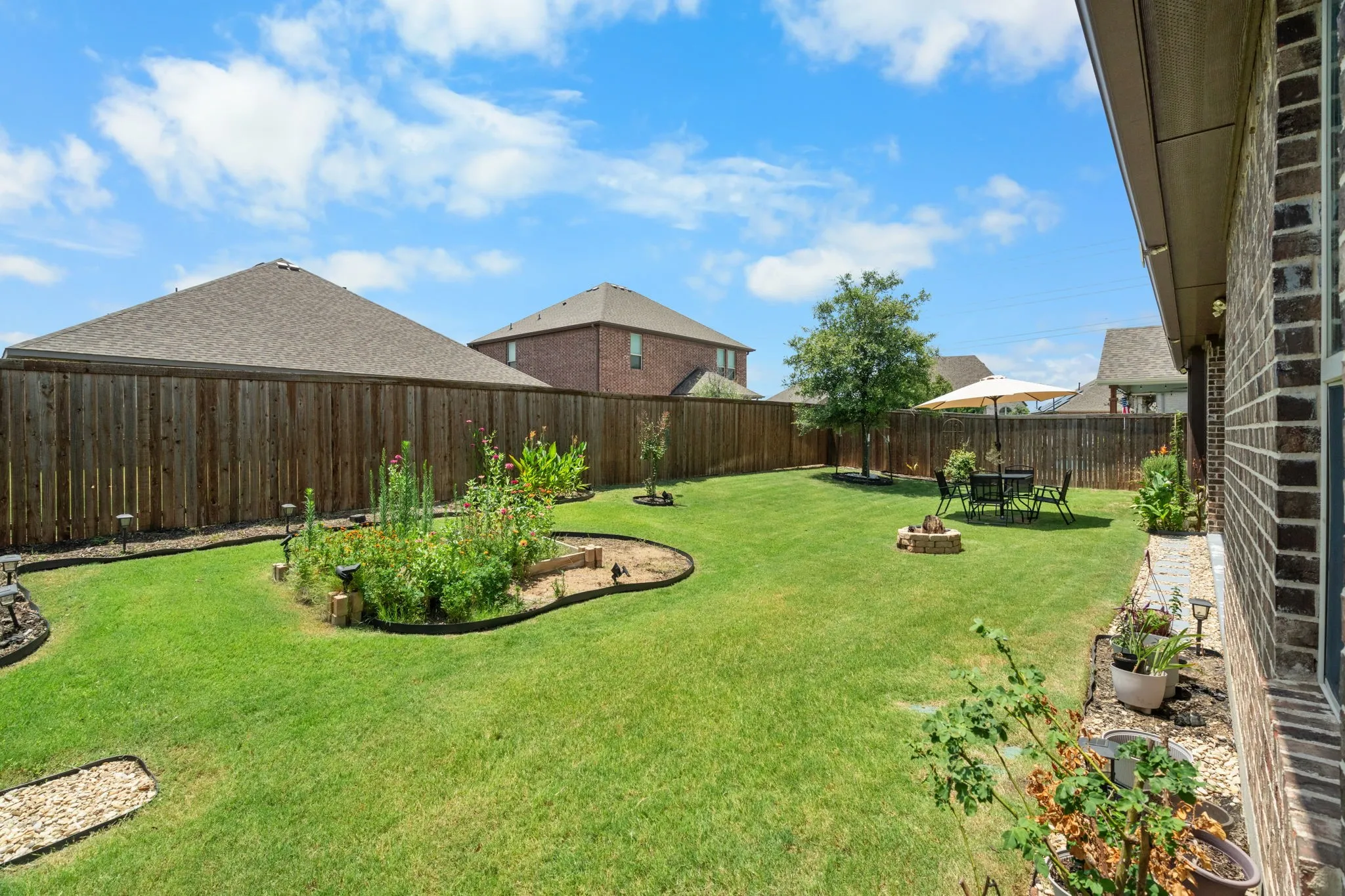 Expansive backyard featuring a manicured lawn, a raised garden bed, a wooden privacy fence, and a brick exterior with a covered patio