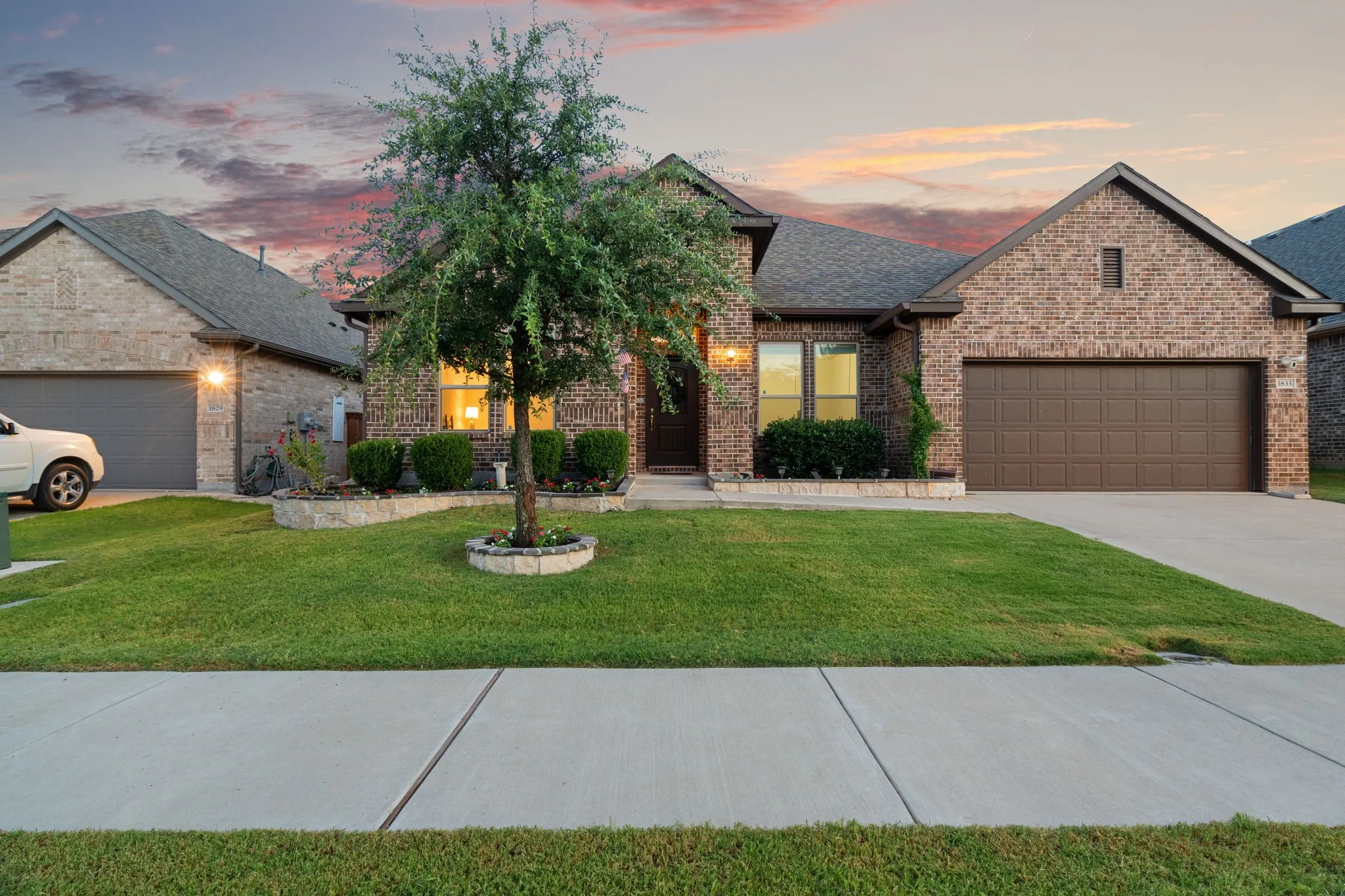 Brick veneer exterior with a dark shingle roof, attached two-car garage, concrete driveway, and a manicured front lawn featuring a stone-edged planter bed