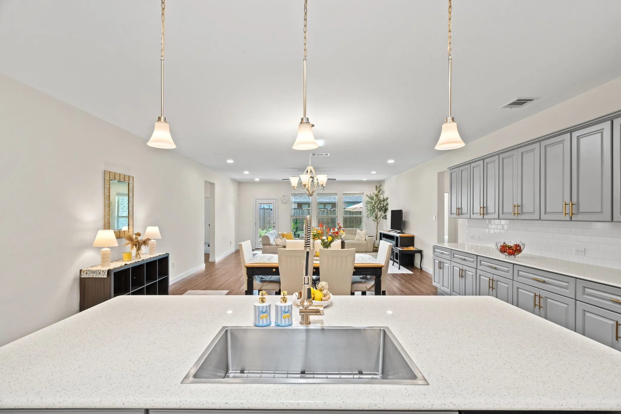 Kitchen island with an undermount stainless steel sink, quartz countertops, gray shaker cabinetry with gold hardware, a white subway tile backsplash, and three pendant lights