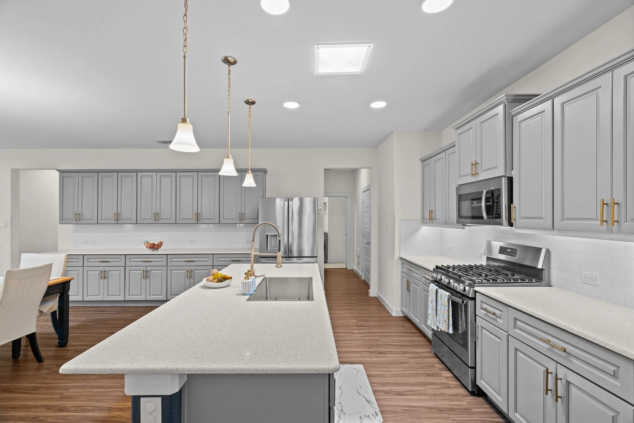 Spacious kitchen featuring an expansive island with an undermount sink, pendant lighting, wood-finish flooring, grey cabinetry with gold-tone hardware, and stainless steel appliances