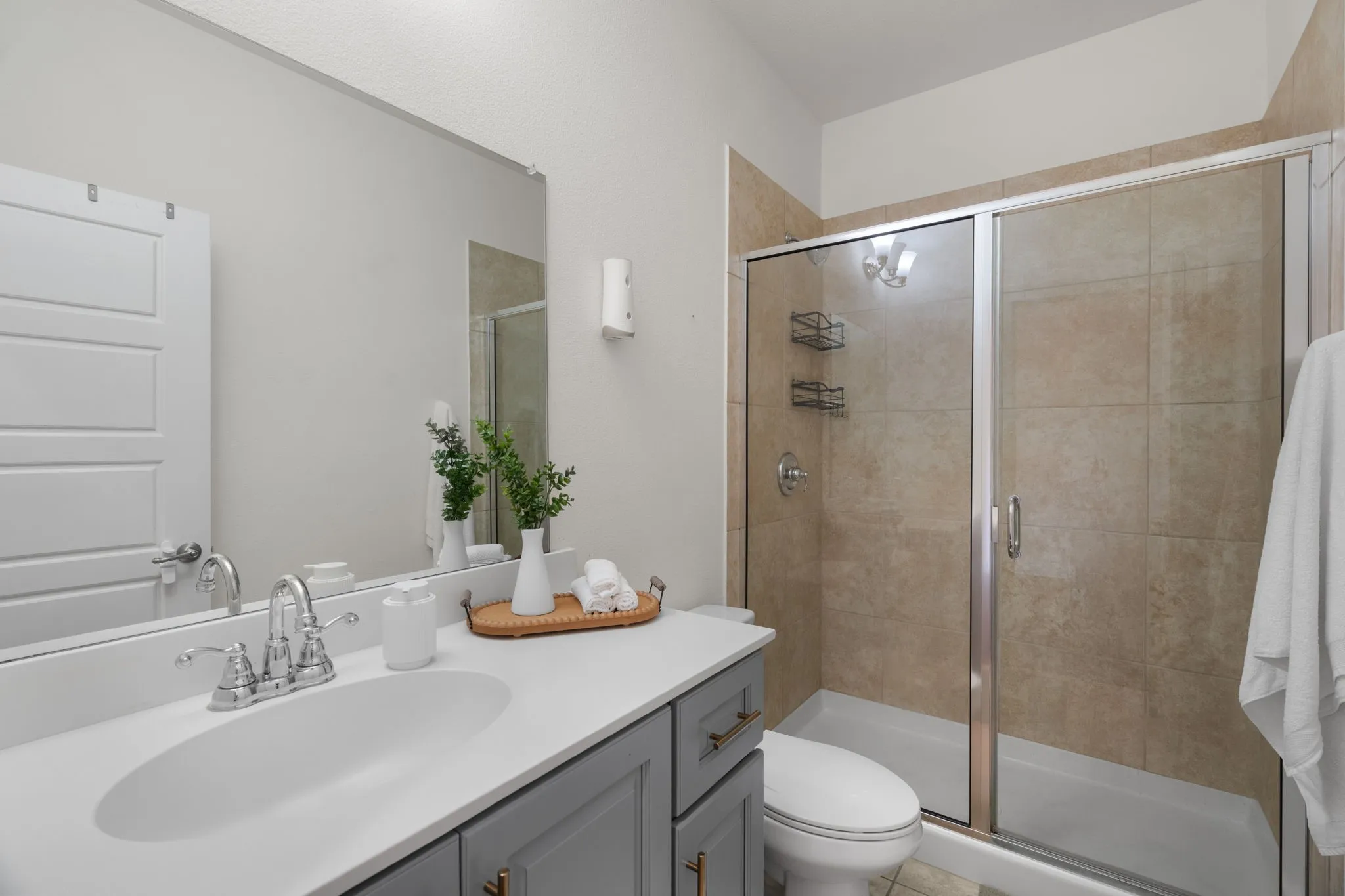 Bathroom featuring a neutral color palette, gray vanity with gold-tone hardware, white countertop with an undermount sink, and a glass-enclosed shower with tiled walls