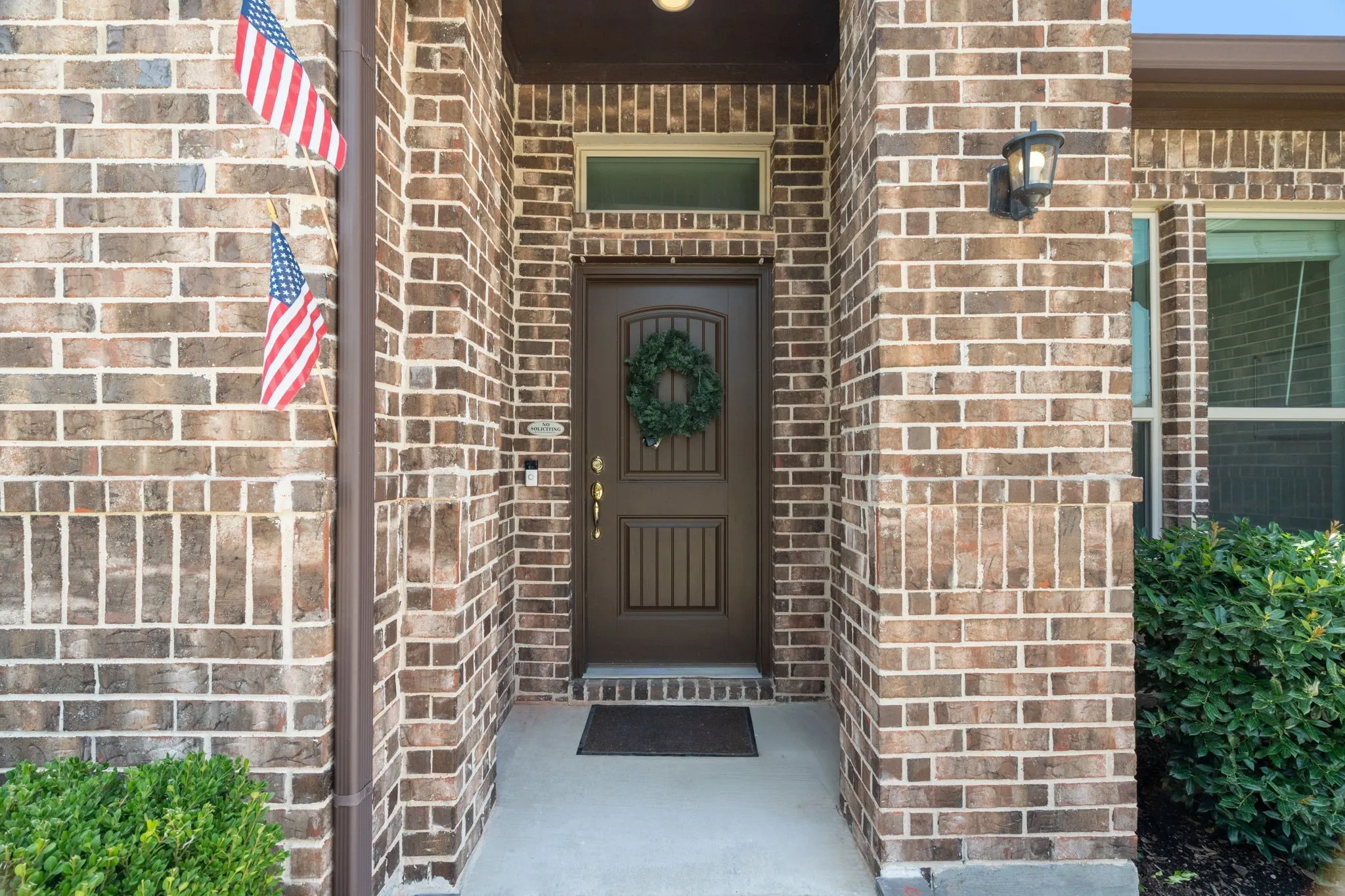 Brick exterior featuring a paneled entry door with transom window, concrete pathway, decorative exterior lighting, and established shrubbery