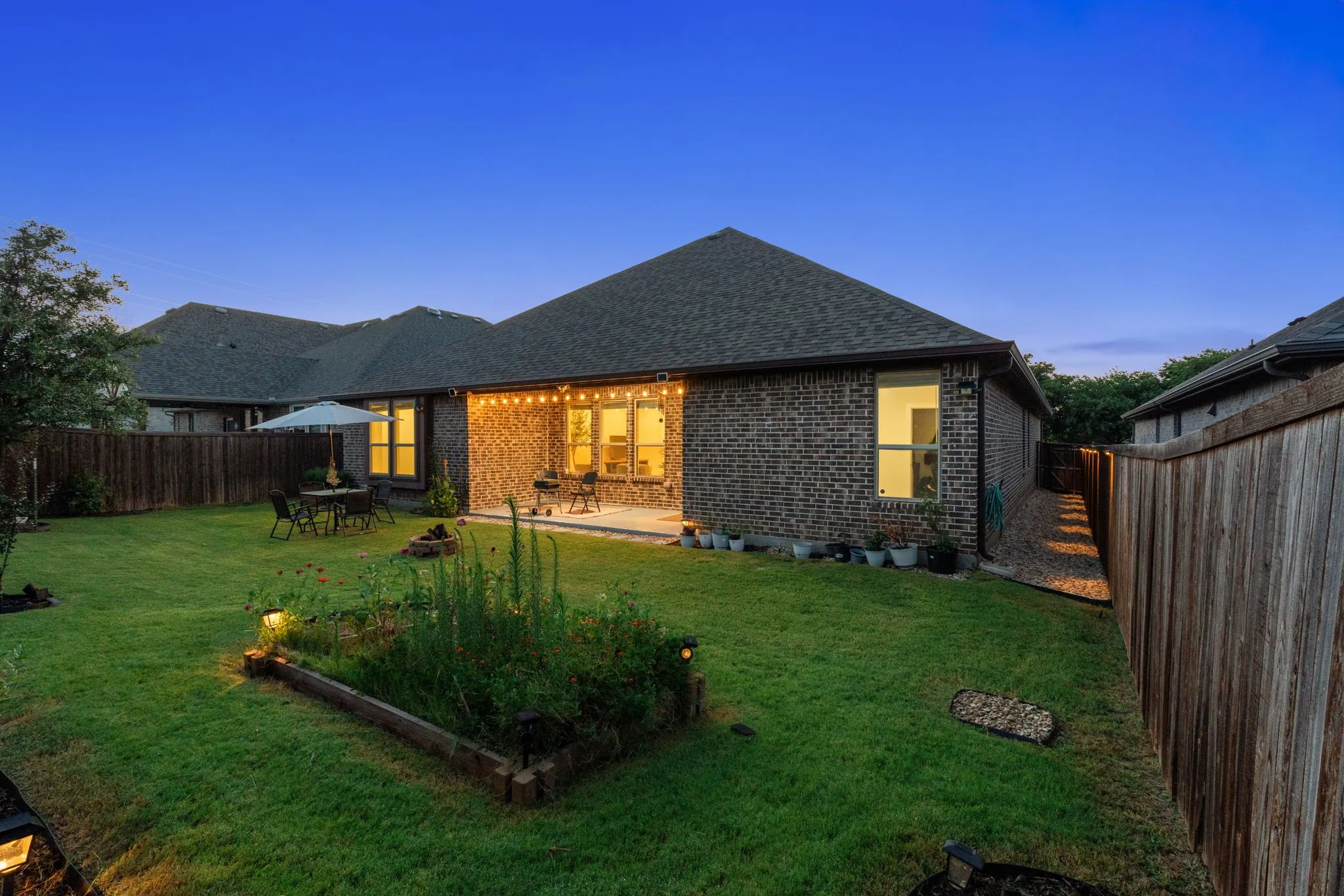 Expansive backyard featuring a manicured lawn, a raised garden bed, and a brick exterior home with a covered patio