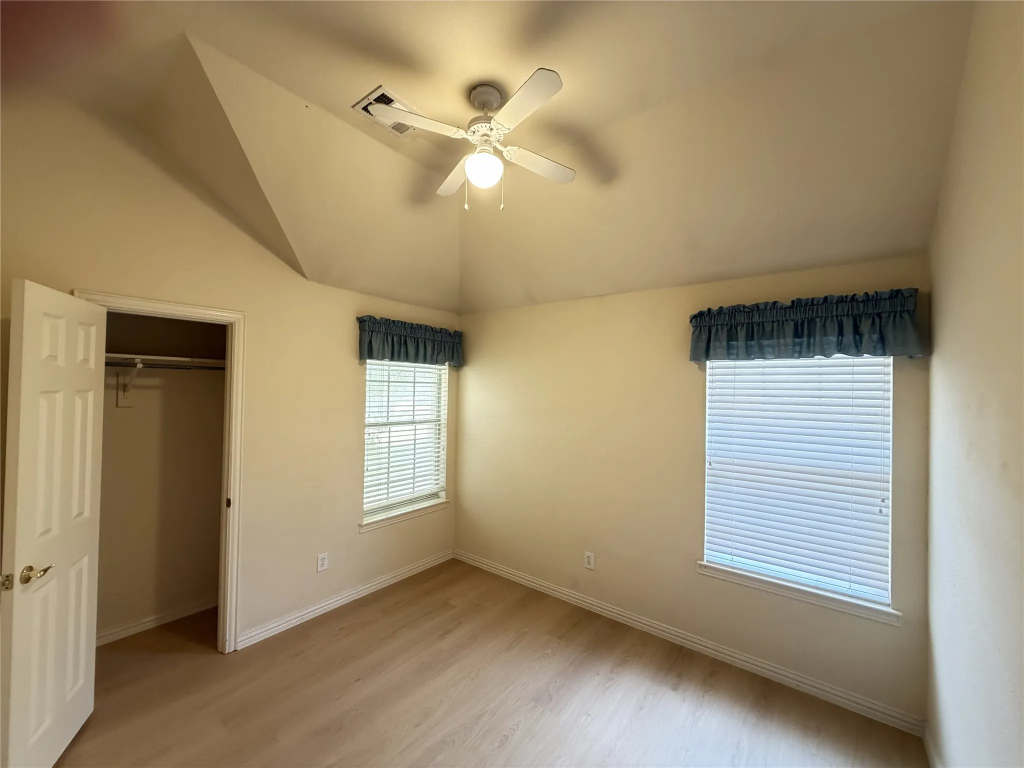 Vaulted ceiling with a ceiling fan, two windows with blinds, wood-finish flooring, and a closet with a built-in rod