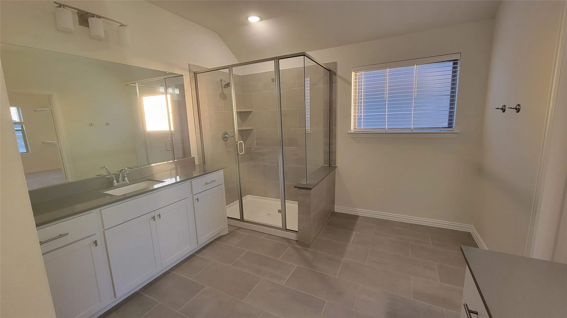 Bathroom featuring a glass-enclosed shower with a built-in bench, a spacious vanity with a single basin sink and stone countertop, white shaker-style cabinetry, and large format tile flooring