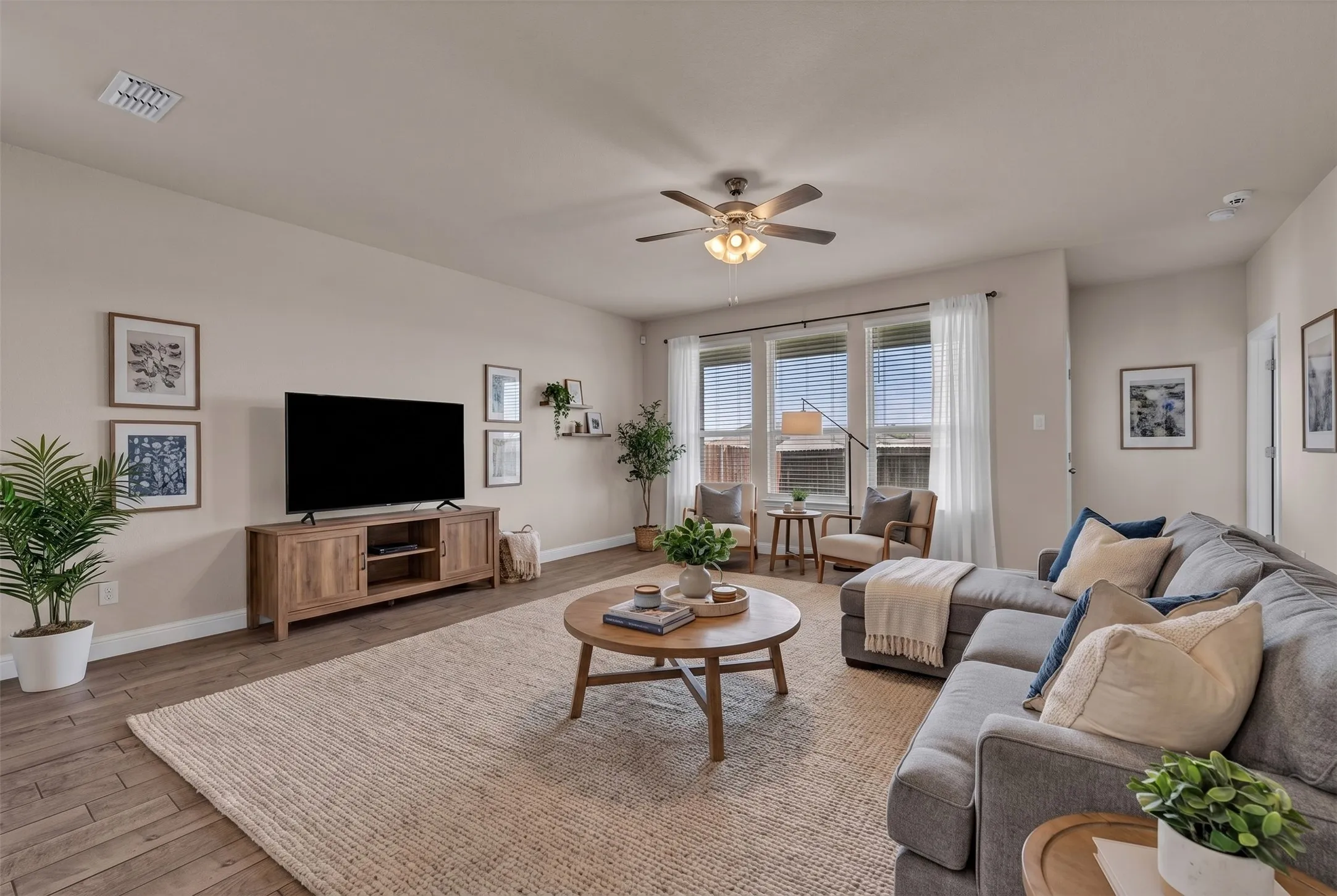 Spacious living area featuring wood-finish flooring, a ceiling fan with integrated lighting, and multiple windows with blinds and sheer curtains