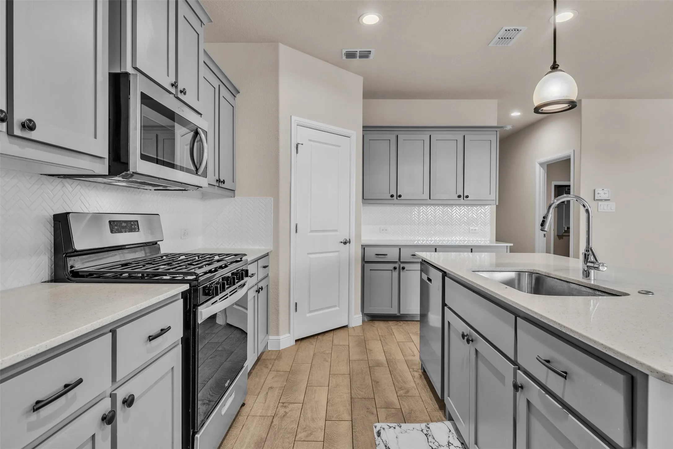 Kitchen featuring light gray cabinetry, stainless steel appliances, light-toned countertops, a white herringbone tile backsplash, and wood-finish flooring