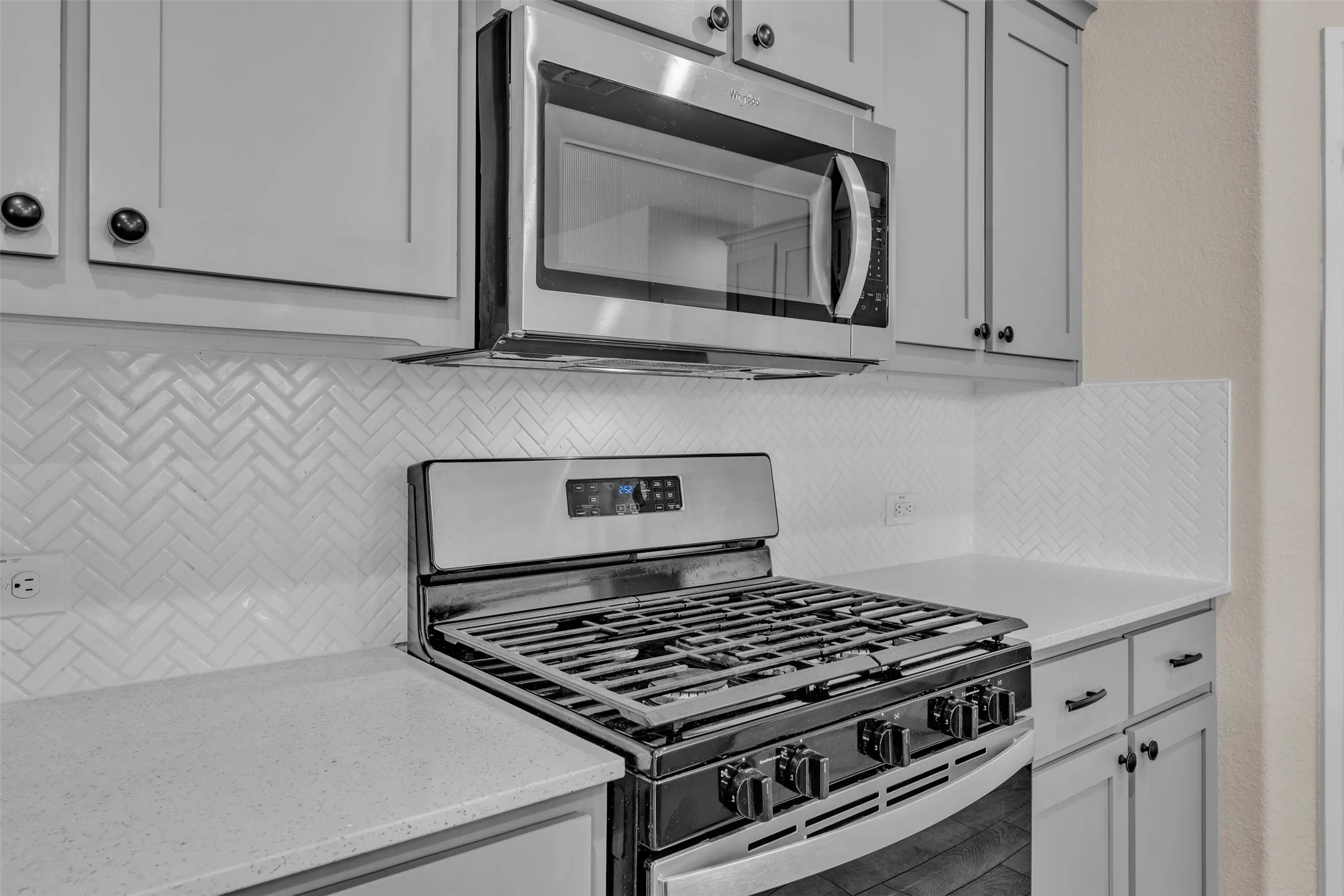 Kitchen featuring a stainless steel gas range, stainless steel microwave, light gray cabinetry with black hardware, white herringbone tile backsplash, and light-toned countertops