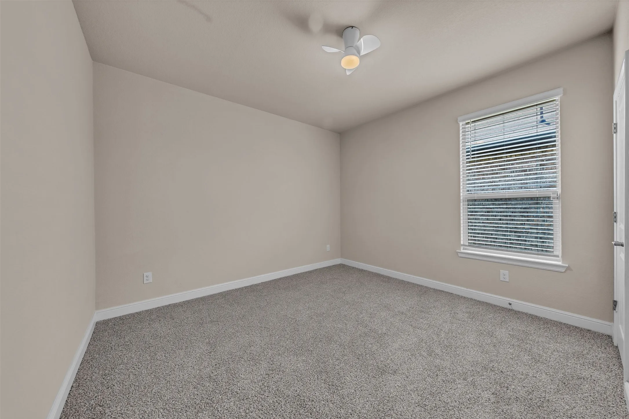 Spacious room featuring neutral paint, gray carpeting, a single window with blinds, white trim, and a ceiling fan with integrated lighting
